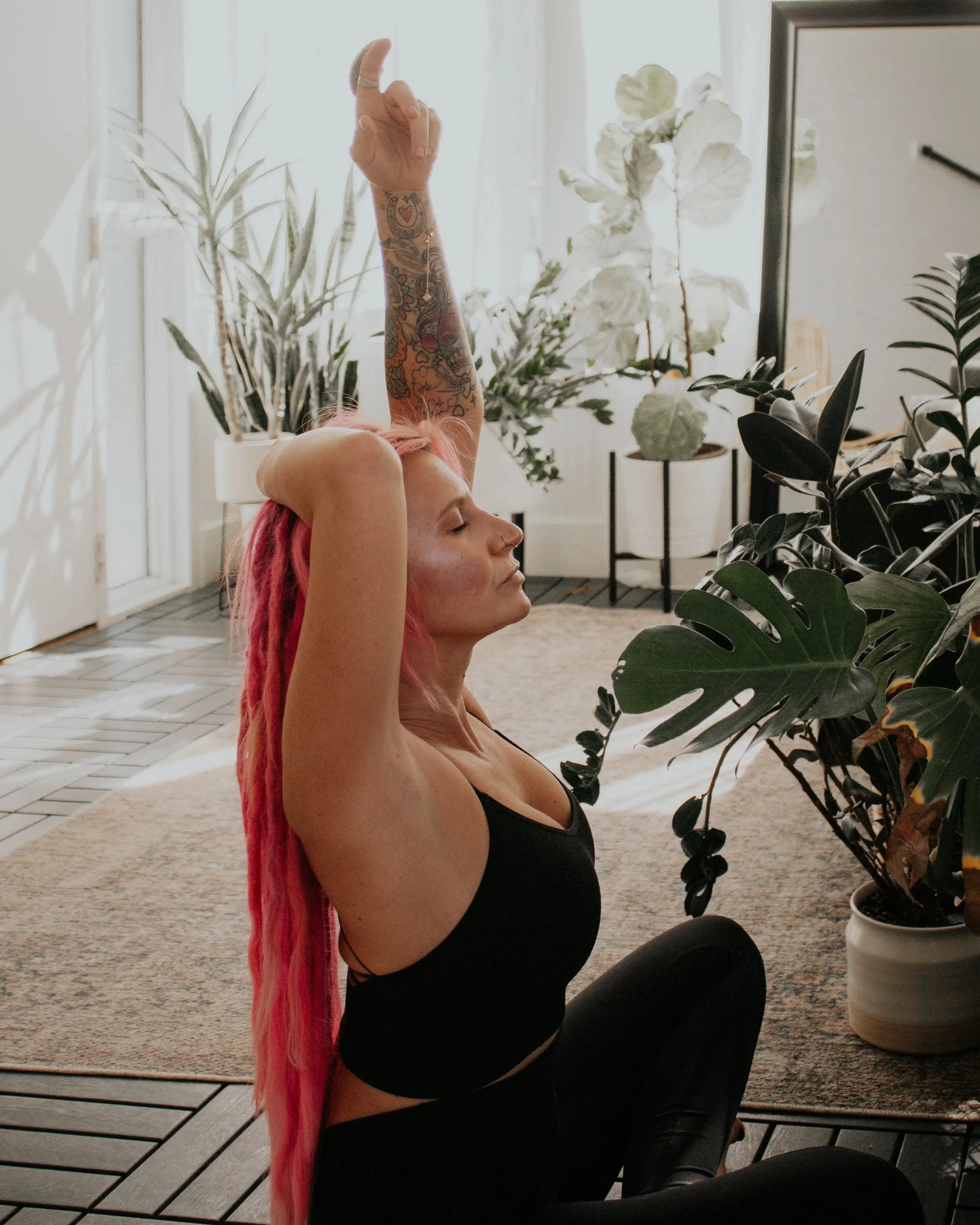 A woman with pink dreadlocks practicing yoga indoors, sitting cross-legged on the floor with her arms raised above her head and eyes closed, surrounded by potted plants.