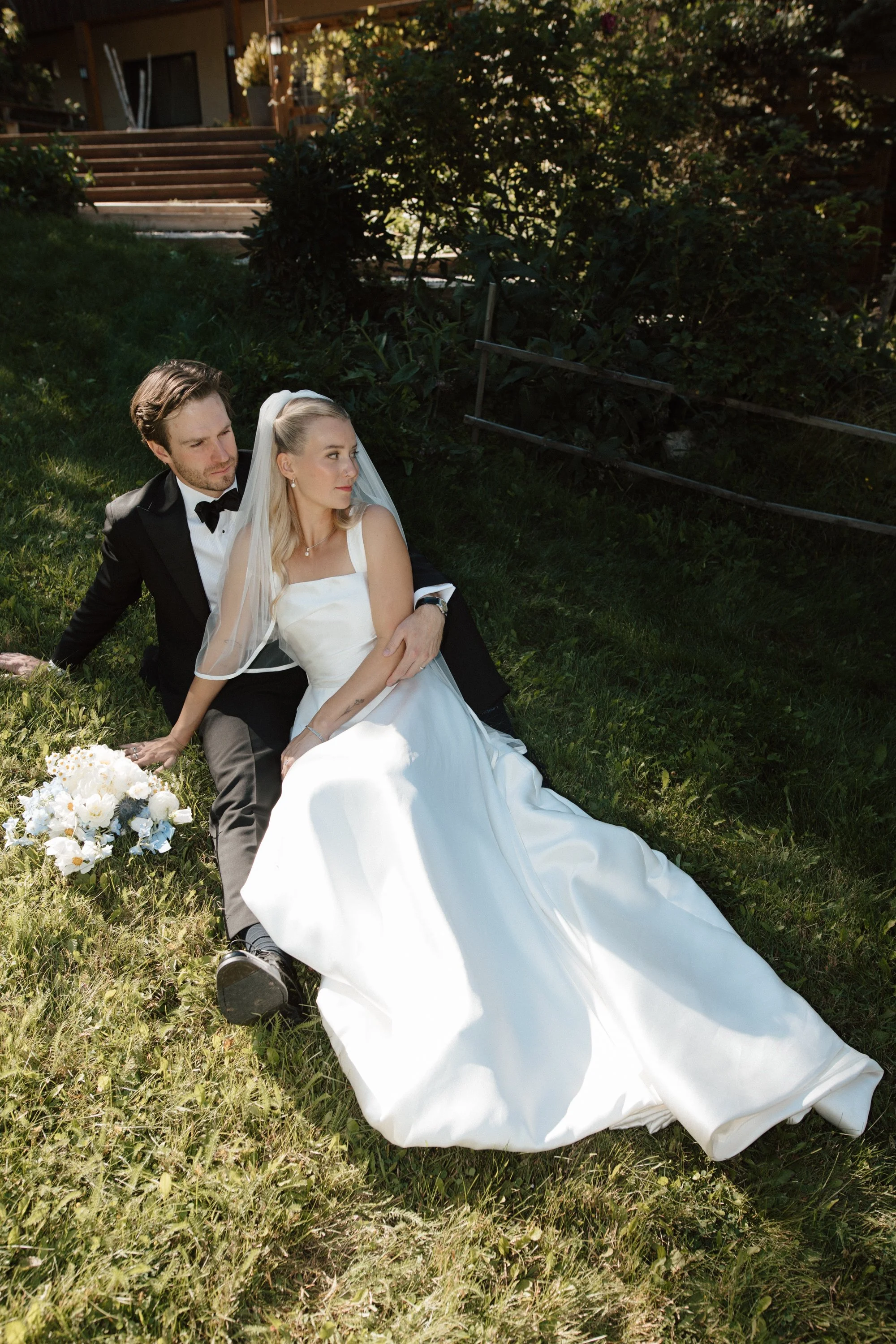 A bride and groom sitting on the grass outdoors, with the bride in a white wedding gown and veil, and the groom in a black tuxedo with a bow tie. The bride has a bouquet of white and blue flowers nearby.
