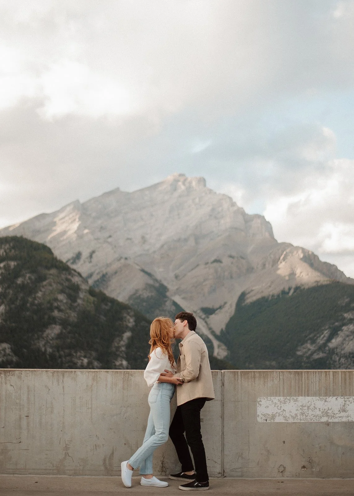 A couple kissing in front of a mountain landscape with clouds in the sky.