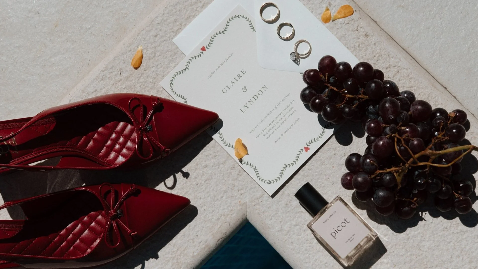 Red high-heeled shoes, wedding rings, a wedding invitation card, a bunch of red grapes, and perfume bottles on a light-colored surface.