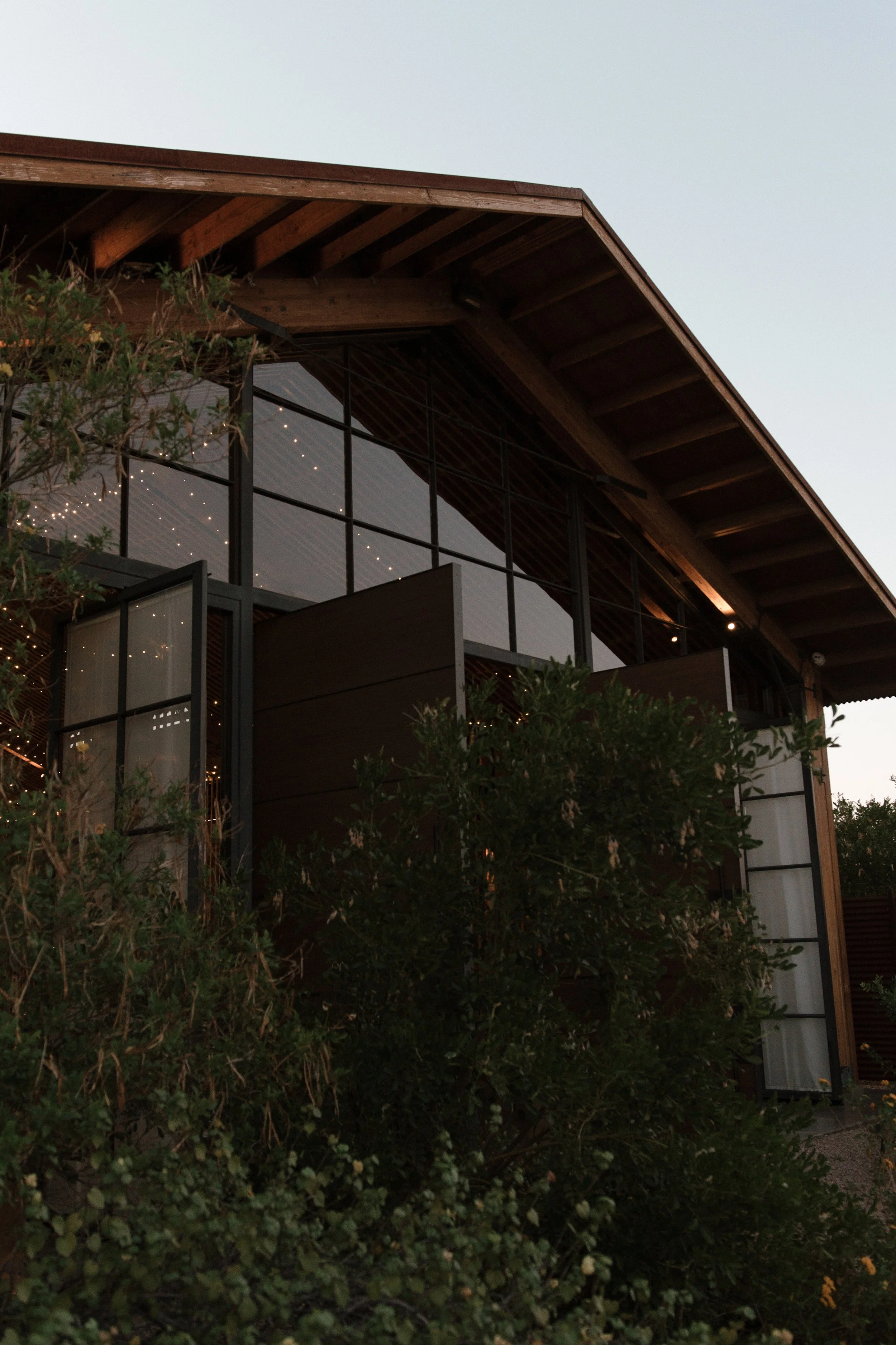 A modern building with large glass windows and a wooden roof structure, surrounded by greenery and trees.