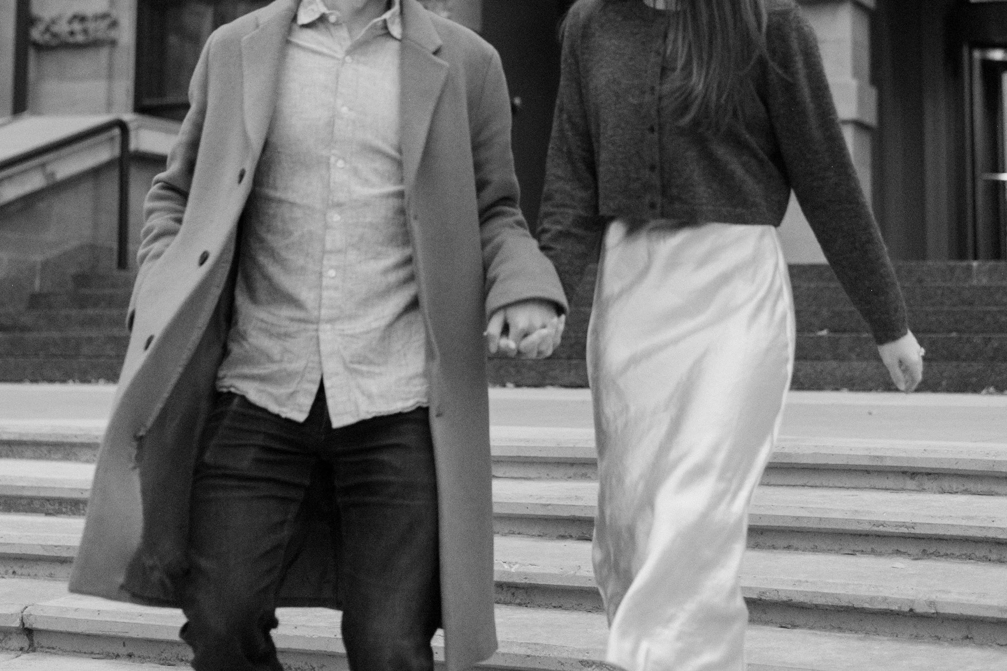A black-and-white photo of two people walking hand-in-hand down a set of stairs. The person on the left is wearing a long coat, shirt, and dark pants. The person on the right is wearing a sweater and light-colored skirt.