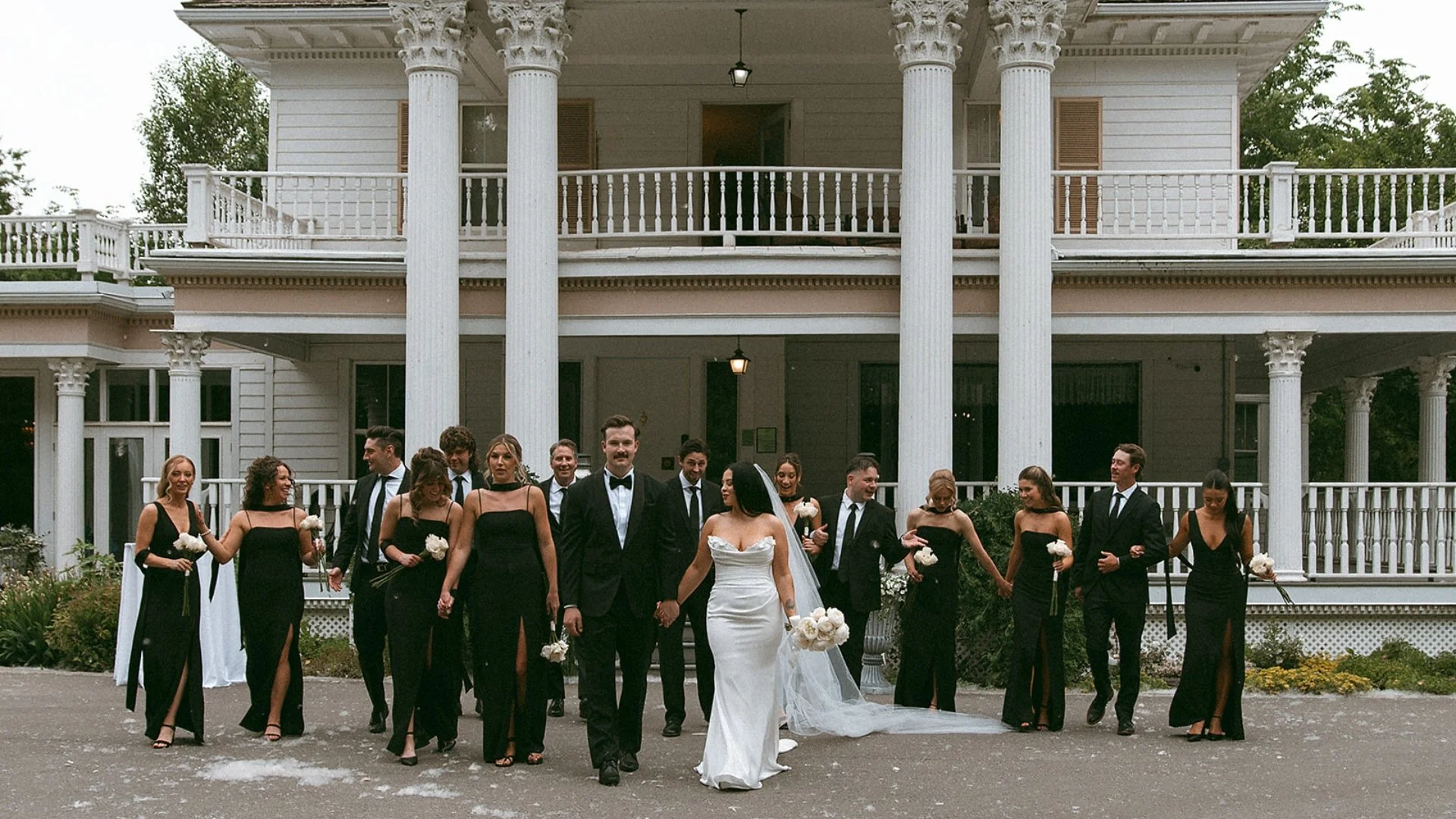 Wedding celebration with bride and groom walking hand in hand, surrounded by bridesmaids and groomsmen, in front of a large white house with columns and balconies.