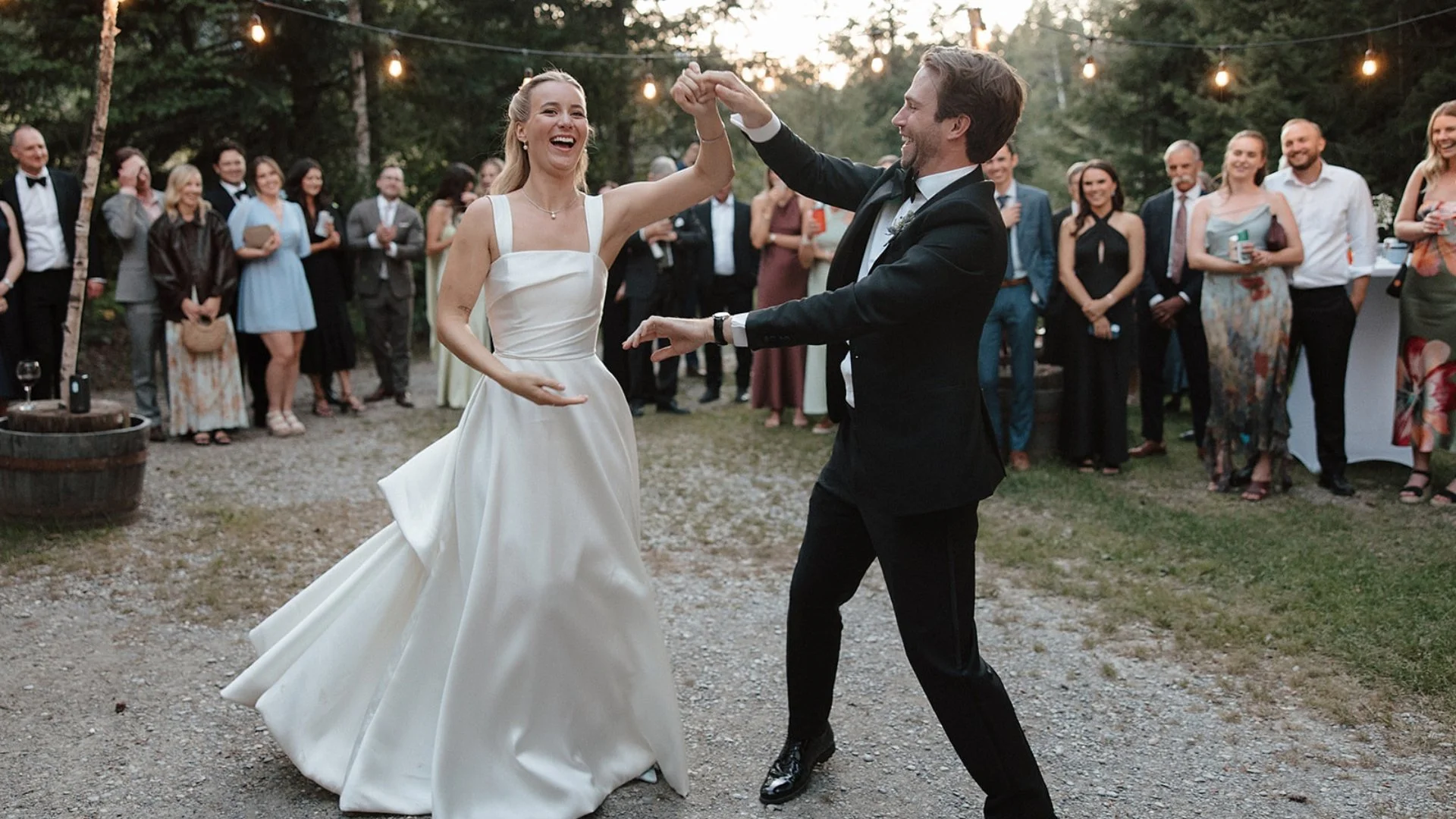 A newlywed couple dances at their outdoor wedding reception surrounded by friends and family. The bride is wearing a white wedding gown, and the groom is in a black tuxedo. Guests are standing around, some holding drinks, smiling and watching the couple dance. String lights hang above the scene in a wooded outdoor setting.