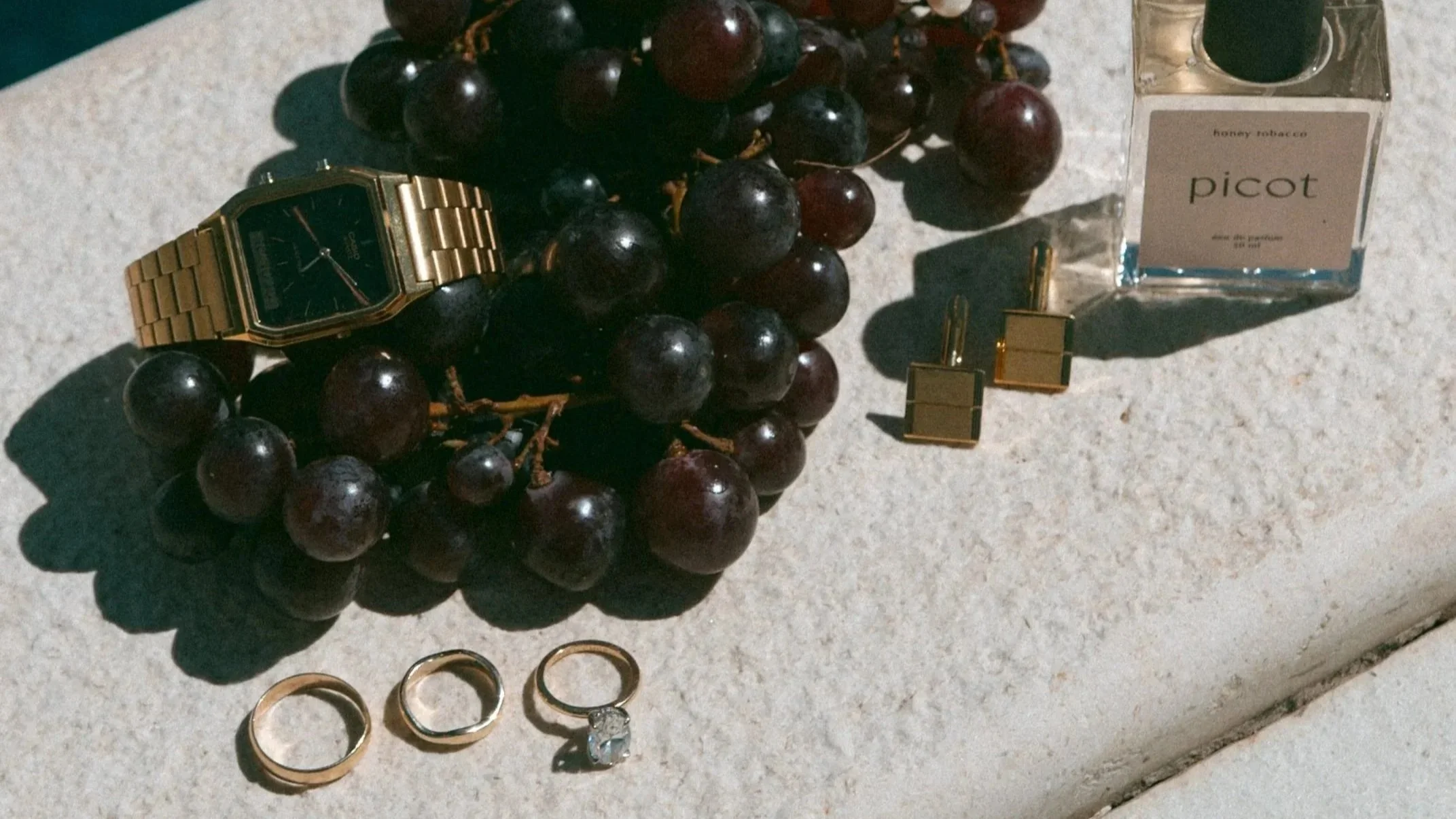 Gold wristwatch, three gold rings, a diamond ring, bunch of grapes, perfume bottle labeled 'picot', and gold cufflinks on a beige surface.