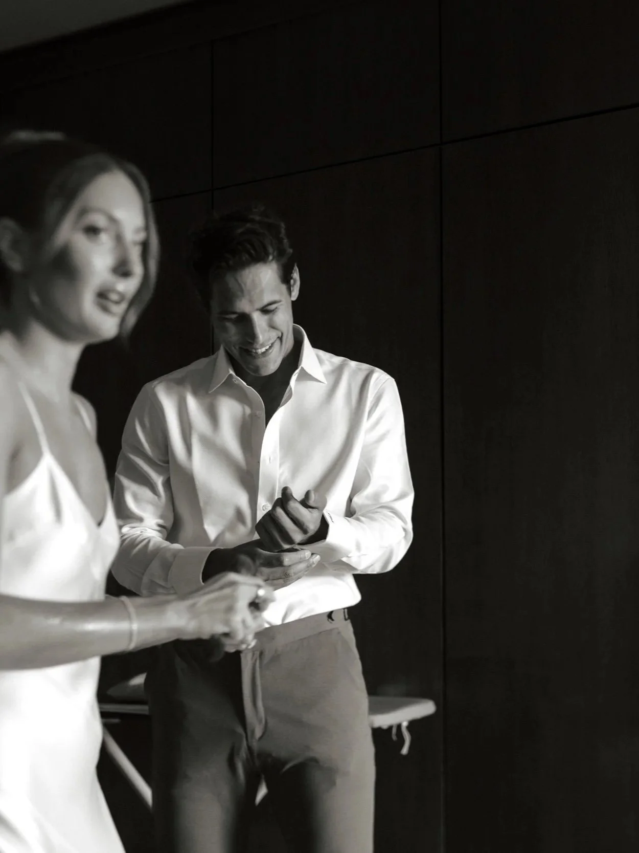 A man and a woman are standing together in a dimly lit room, both holding and looking at their phones. The man is smiling and wearing a white dress shirt, and the woman is slightly blurred, wearing a white sleeveless dress. They are standing against a dark wall, and there is a small table or stool in the background.