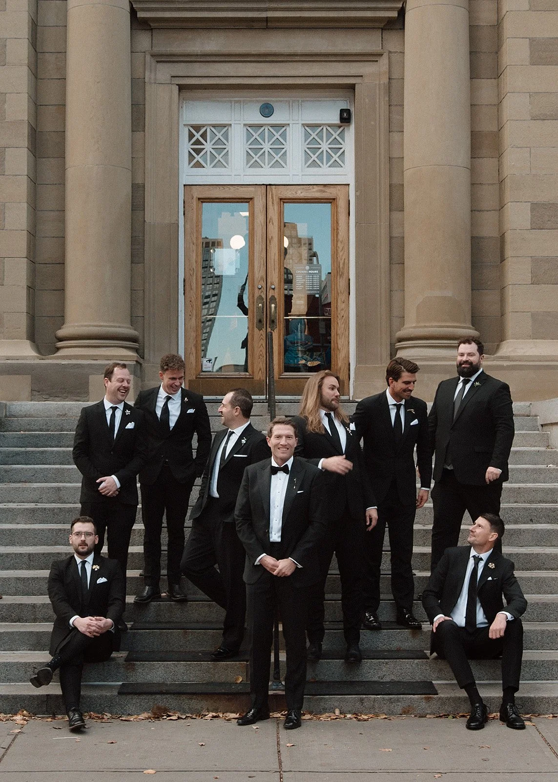 A group of men in tuxedos posing on the steps of a building with large columns and wooden doors.