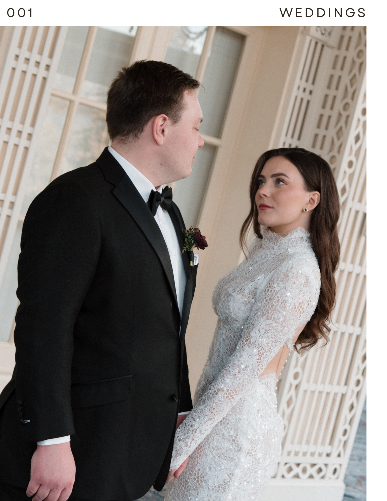A couple dressed in formal wedding attire holding hands and gazing into each other's eyes indoors near a decorative white wooden wall.