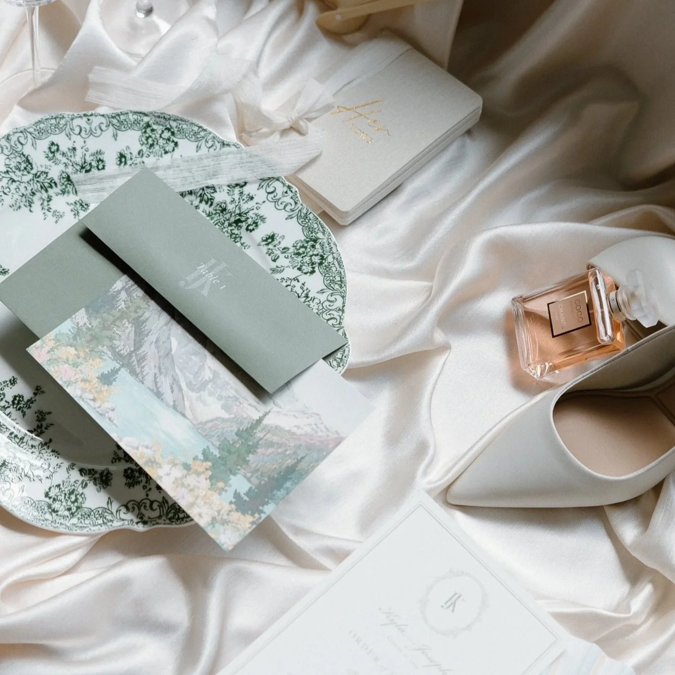 A flat lay display of a floral-patterned tray with wedding details, including a grey envelope with the word 'Thee,' a scenic card of mountains and a lake, a small white book with gold lettering, a bottle of pink perfume labeled 'Coco', white shoes, and a white robe, all arranged on cream-colored satin fabric.