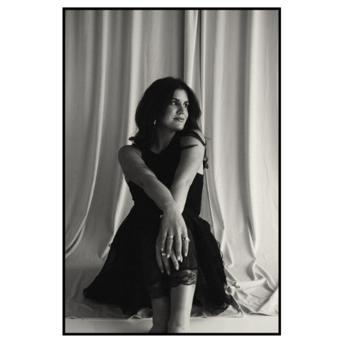 A woman sitting on the floor in front of curtains, wearing a black dress, looking to her right with a serene expression.