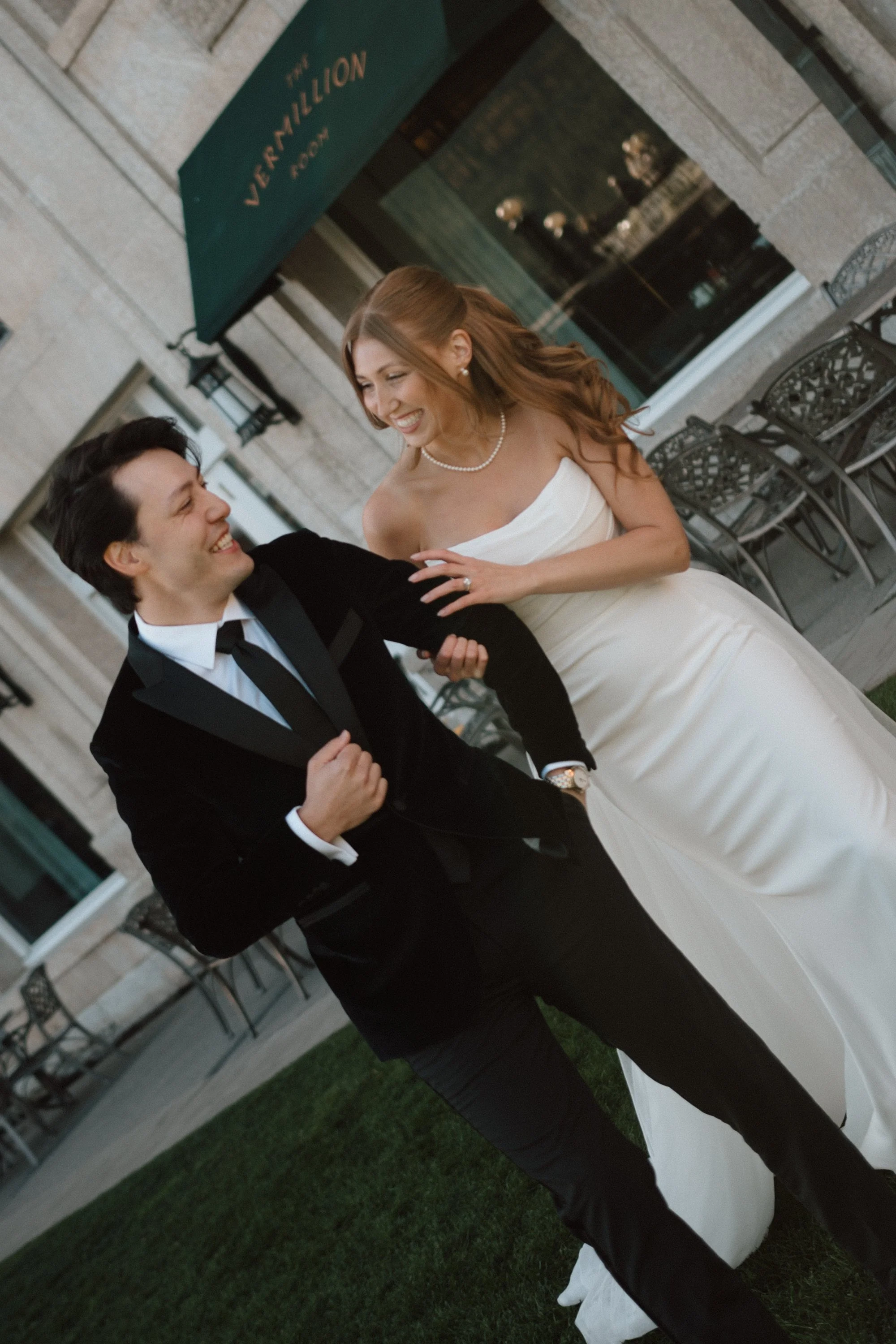 A smiling couple, dressed in formal wedding attire, standing close together outside a building with a sign that reads 'The Vern Miller Room.' The woman is wearing a strapless white wedding gown and pearl jewelry, while the man is in a black tuxedo with a bow tie. They are holding hands, and the woman has a ring on her finger, indicating a wedding celebration.