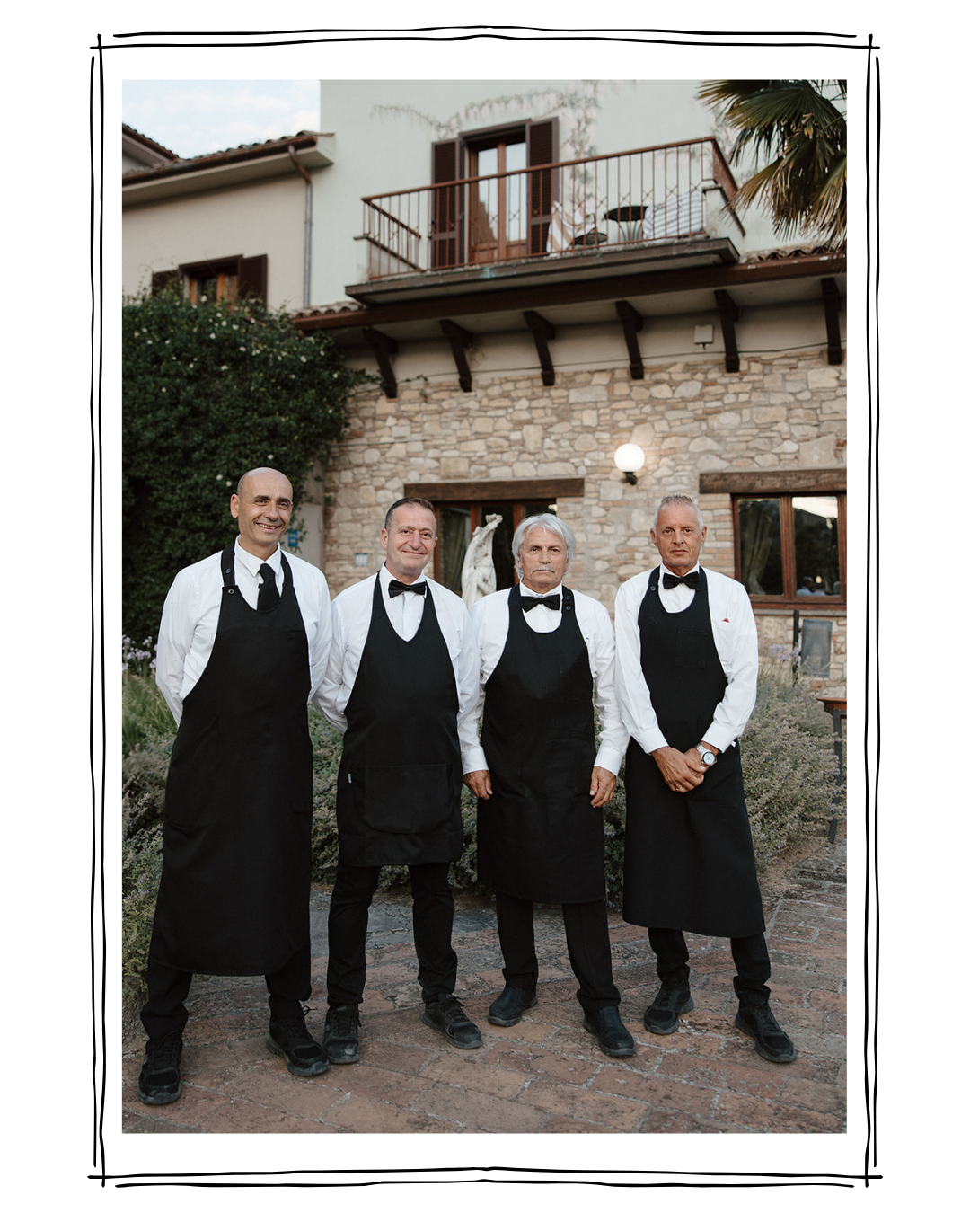 Four male waiters in black aprons and white shirts standing in front of a stone building during daytime.
