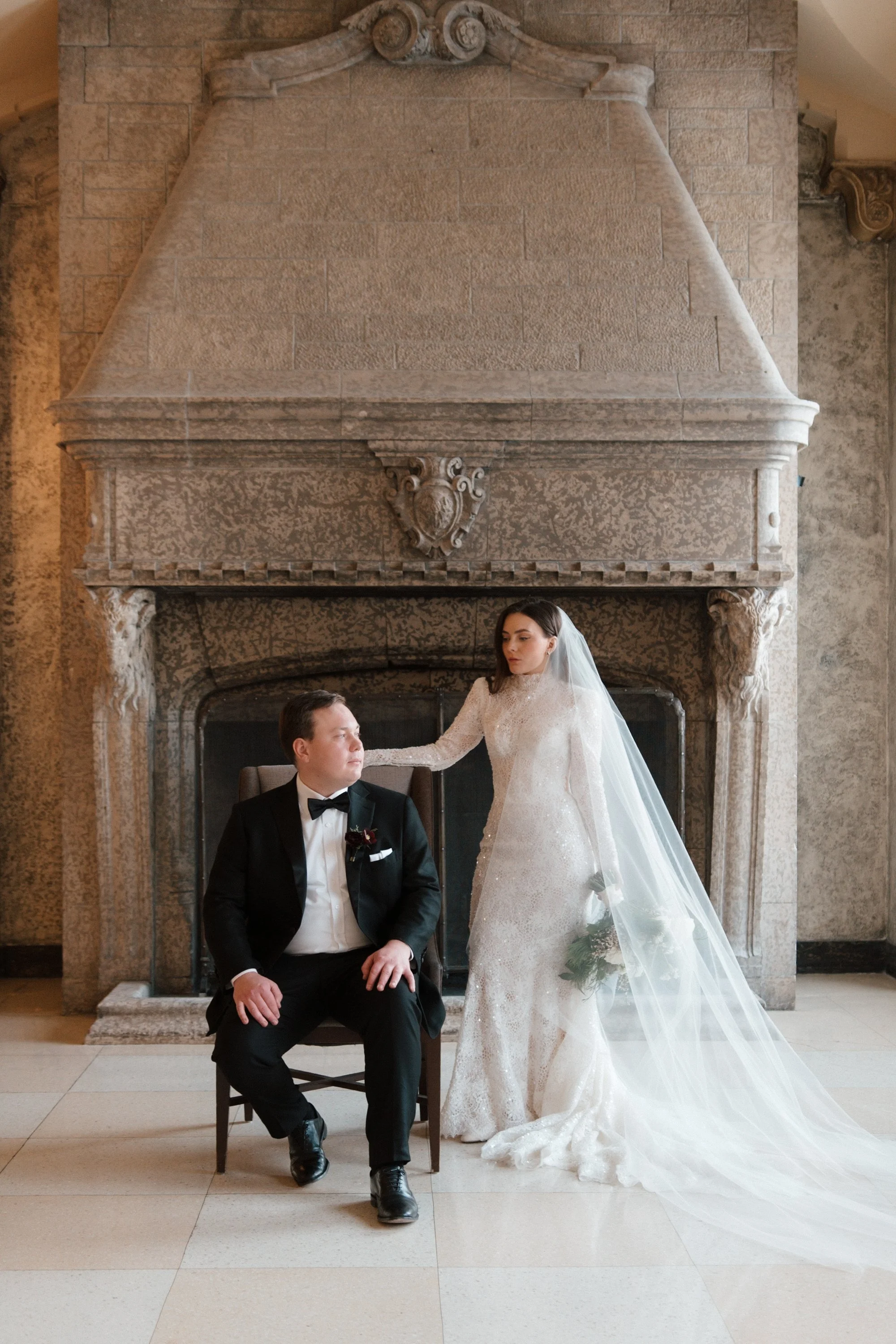 A bride and groom in wedding attire pose in front of a large ornate stone fireplace with a carved crest. The groom, sitting in a chair, wears a black tuxedo with a bow tie, and the bride, standing with her hand on his shoulder, wears a long white wed