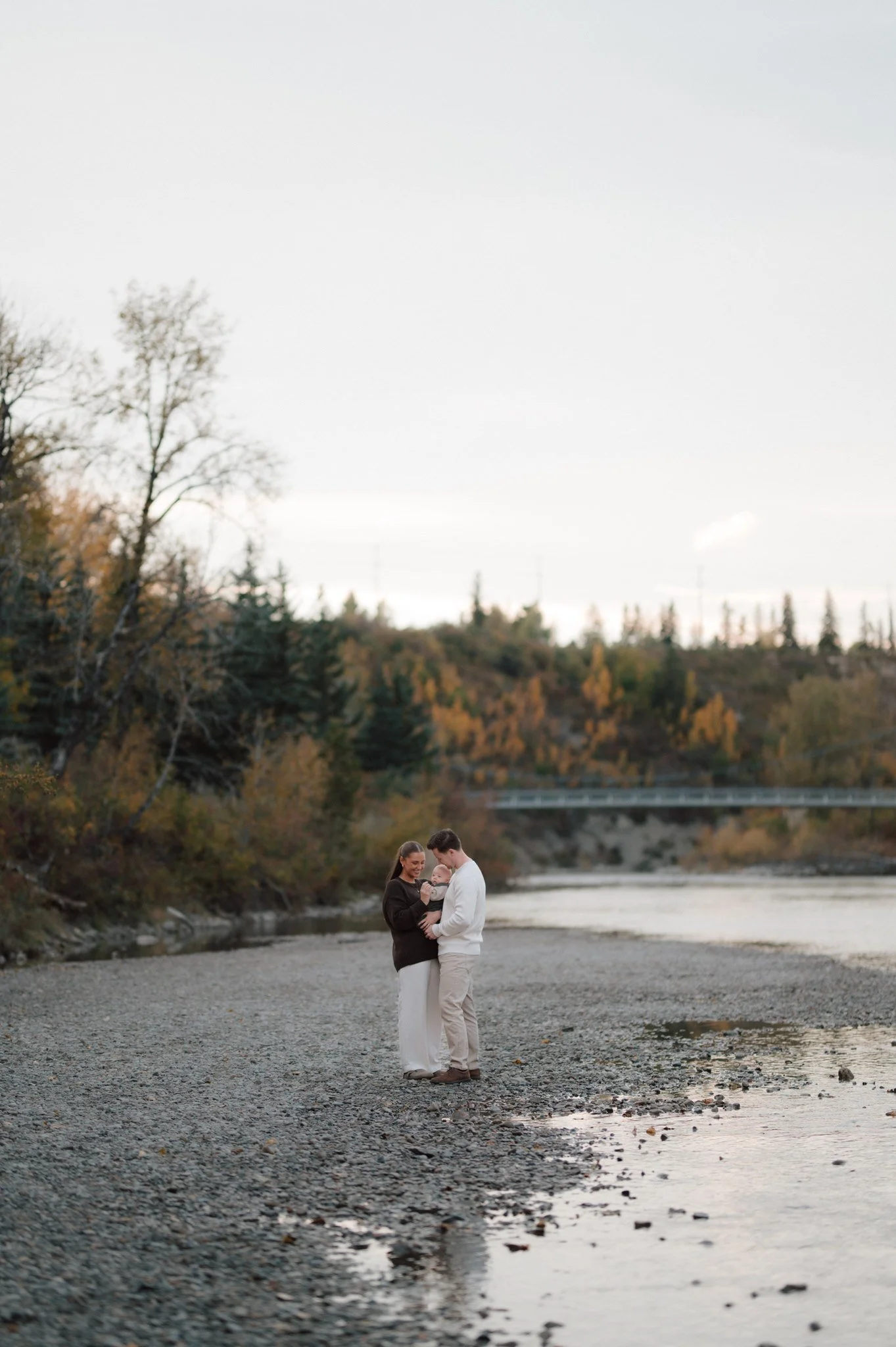 A couple and a baby standing on a rocky riverside, with trees and a bridge in the background during autumn.