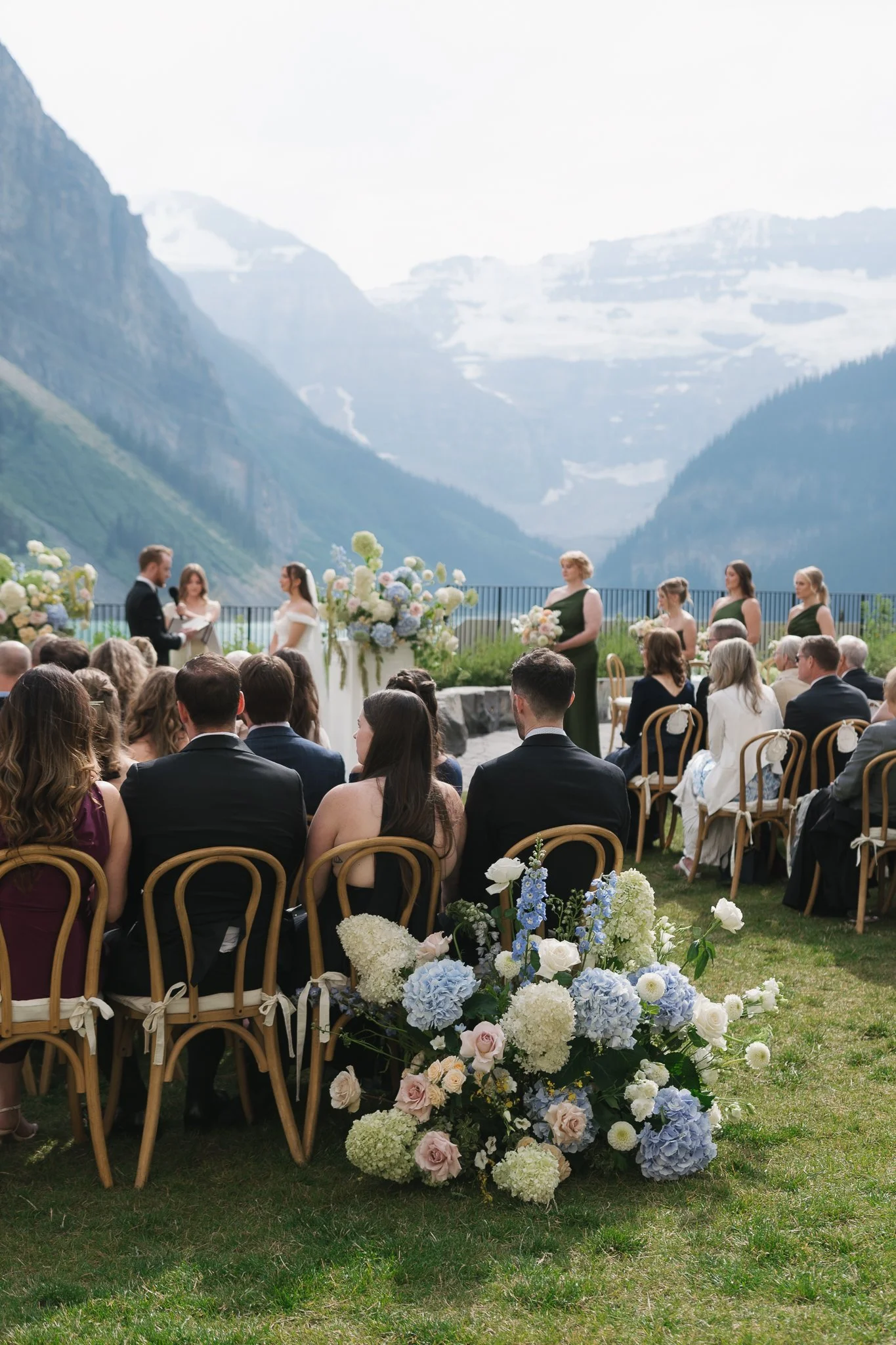 A wedding ceremony taking place outdoors with mountains in the background, featuring the bride and groom exchanging vows, surrounded by floral arrangements, with guests seated on chairs and a large bouquet of flowers in the foreground.