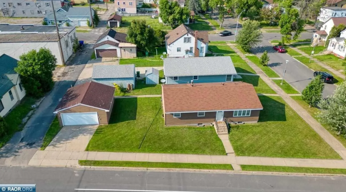 An aerial view of a residential neighborhood featuring a single-story house with a red roof, a front porch, and a well-maintained lawn, along with a detached garage and various other houses and green spaces.