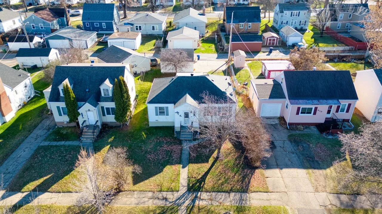 Aerial view of a residential neighborhood showing a row of small single-family homes with detached garages, pitched roofs, and tree-lined yards, captured from above on a clear day.