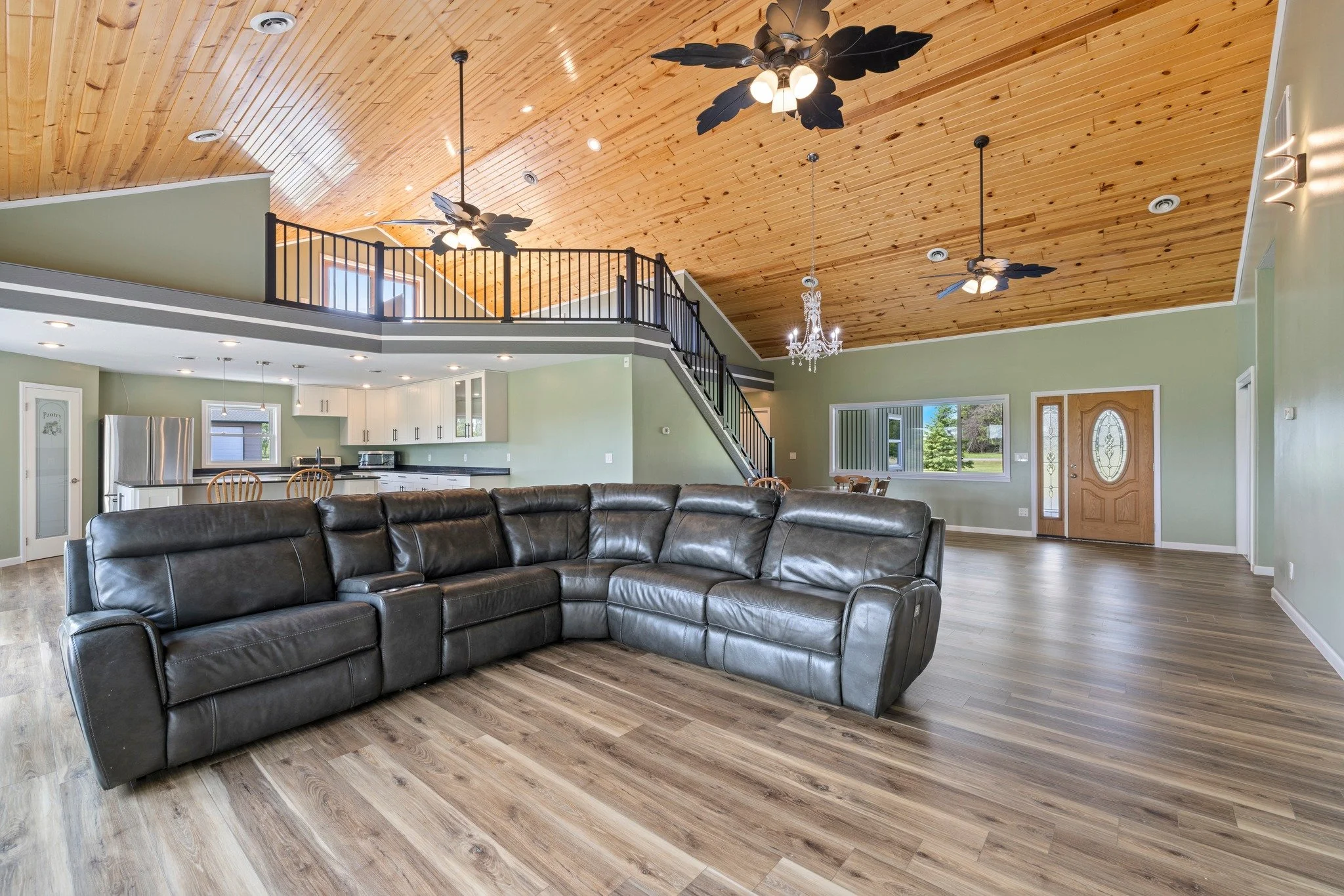 Spacious open living room with black leather sectional sofa, light green walls, wooden floor, and a vaulted wooden ceiling with ceiling fans and lights. Behind the sofa is a kitchen with white cabinets and stainless steel appliances, including a refrigerator.