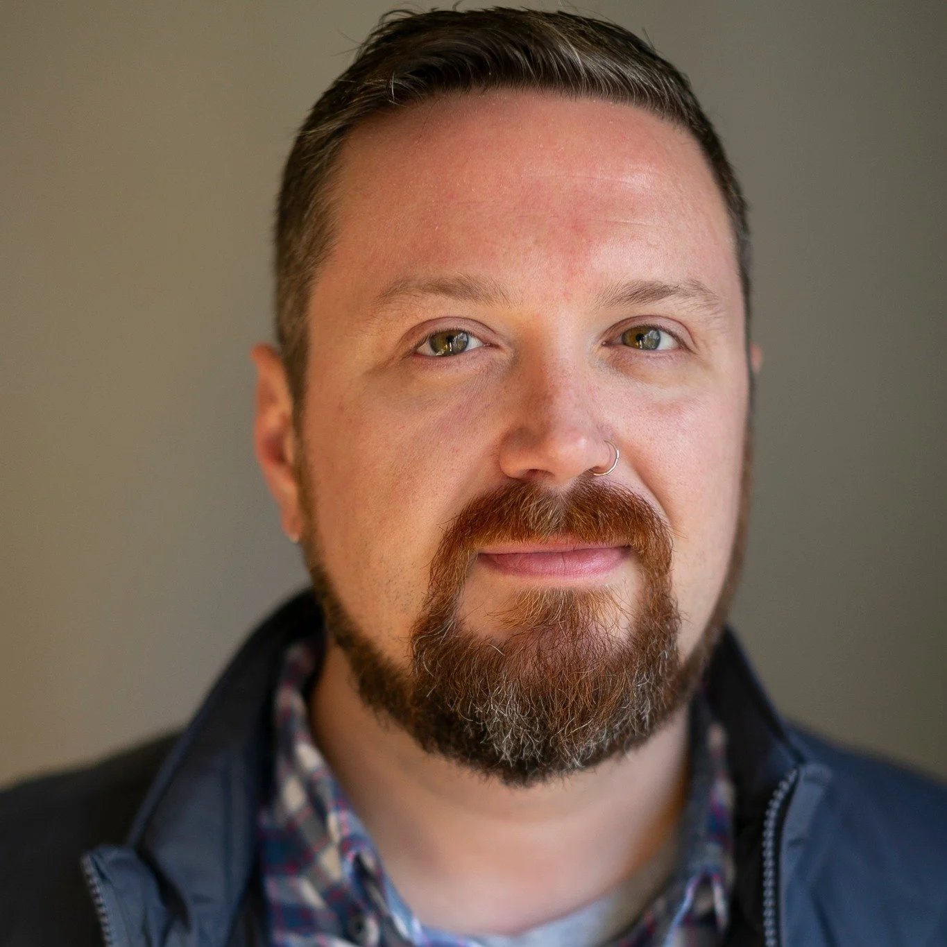 Close-up portrait of a man with short dark hair, a beard, and a nose ring, wearing a black jacket over a plaid shirt, against a neutral background.