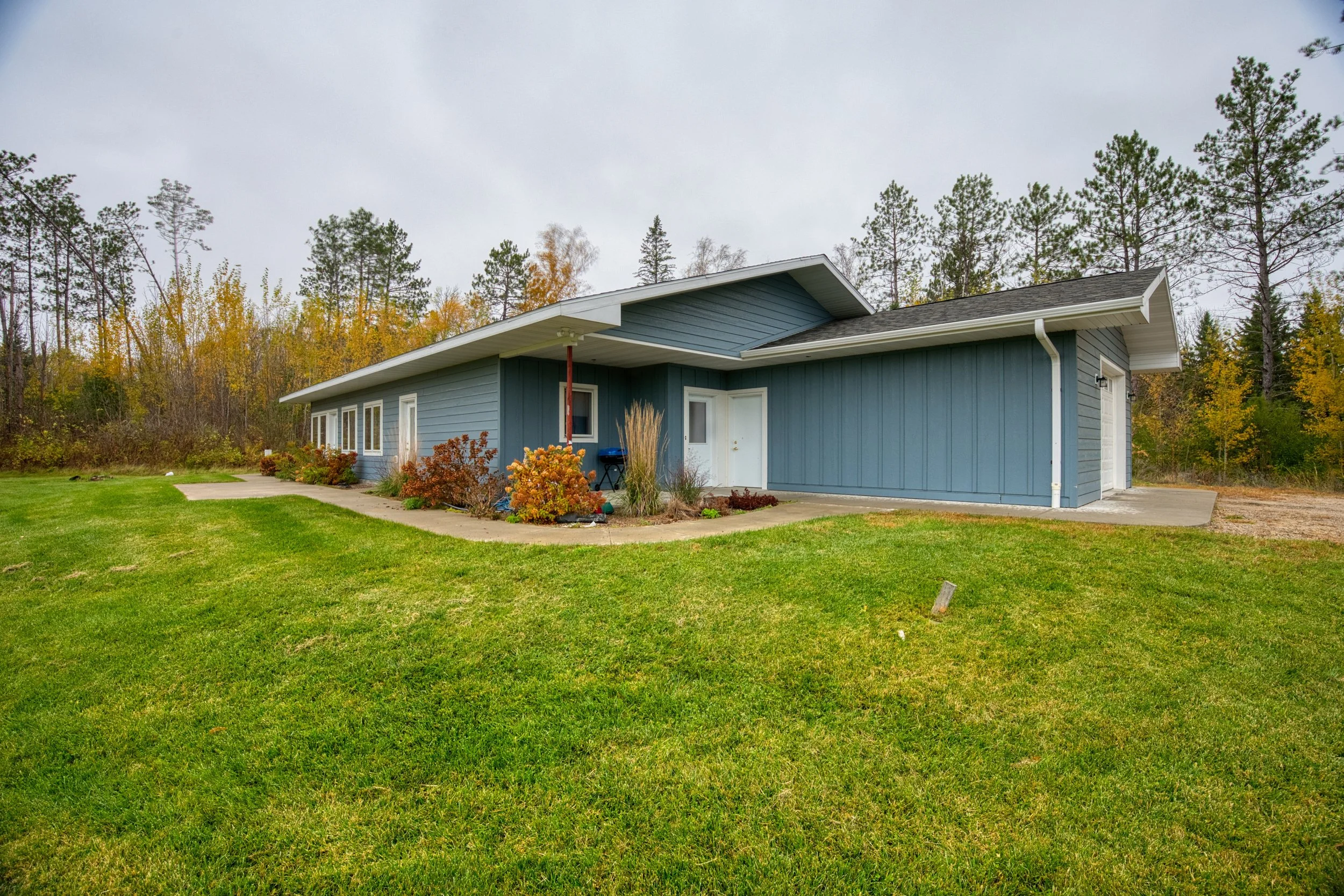 An exterior view of a single-story home with blue-gray siding and a dark blue/charcoal roof. The house has a low-slope, modern roofline and an attached one-stall garage on the right side. White trim accents the roofline, windows, and doors.