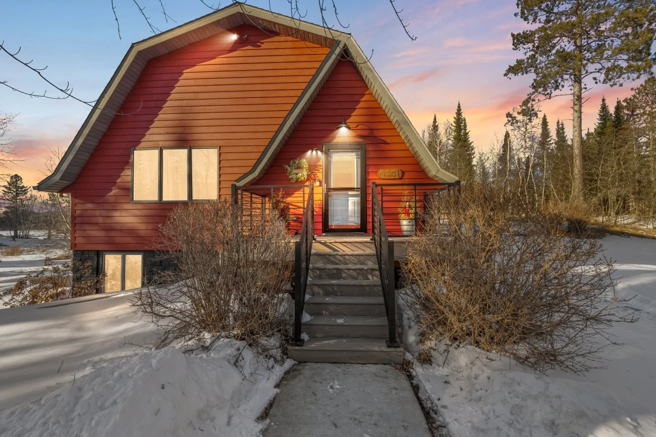 A red house with black railings on a small set of stairs leading to a front door, situated in a snowy landscape with trees and a colorful sunset sky in the background.
