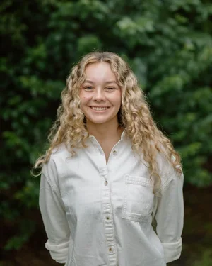 Young woman with curly blond hair smiling outdoors in front of green leafy trees.