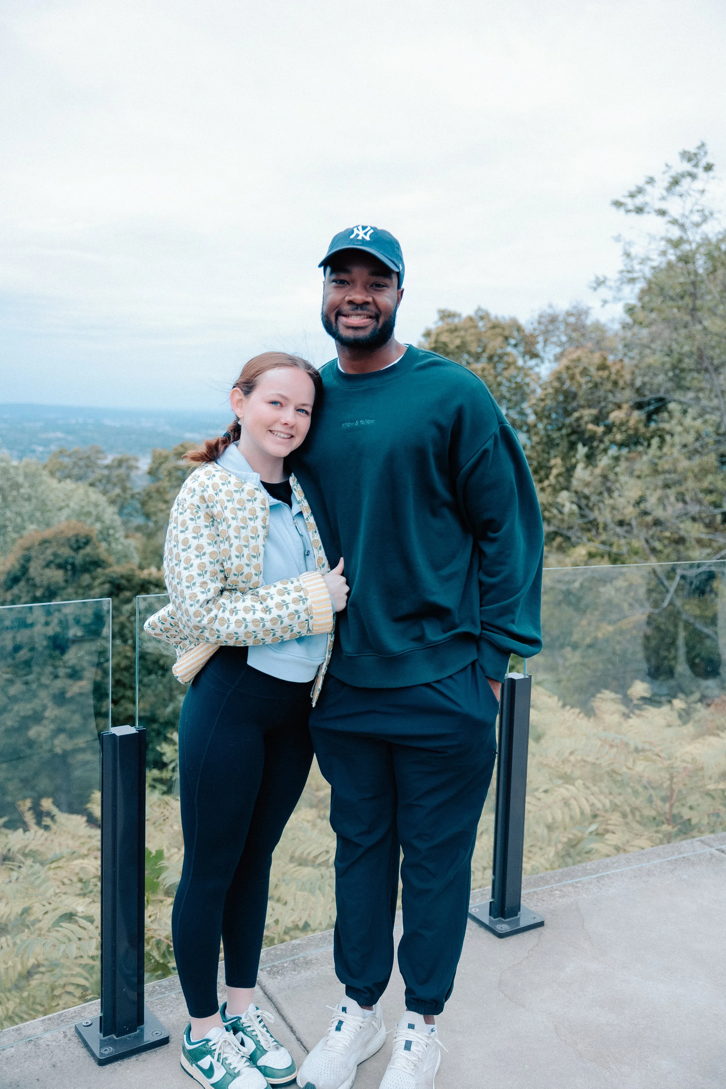 A young woman and a man posing together outdoors on a balcony with a view of trees and a cloudy sky in the background.