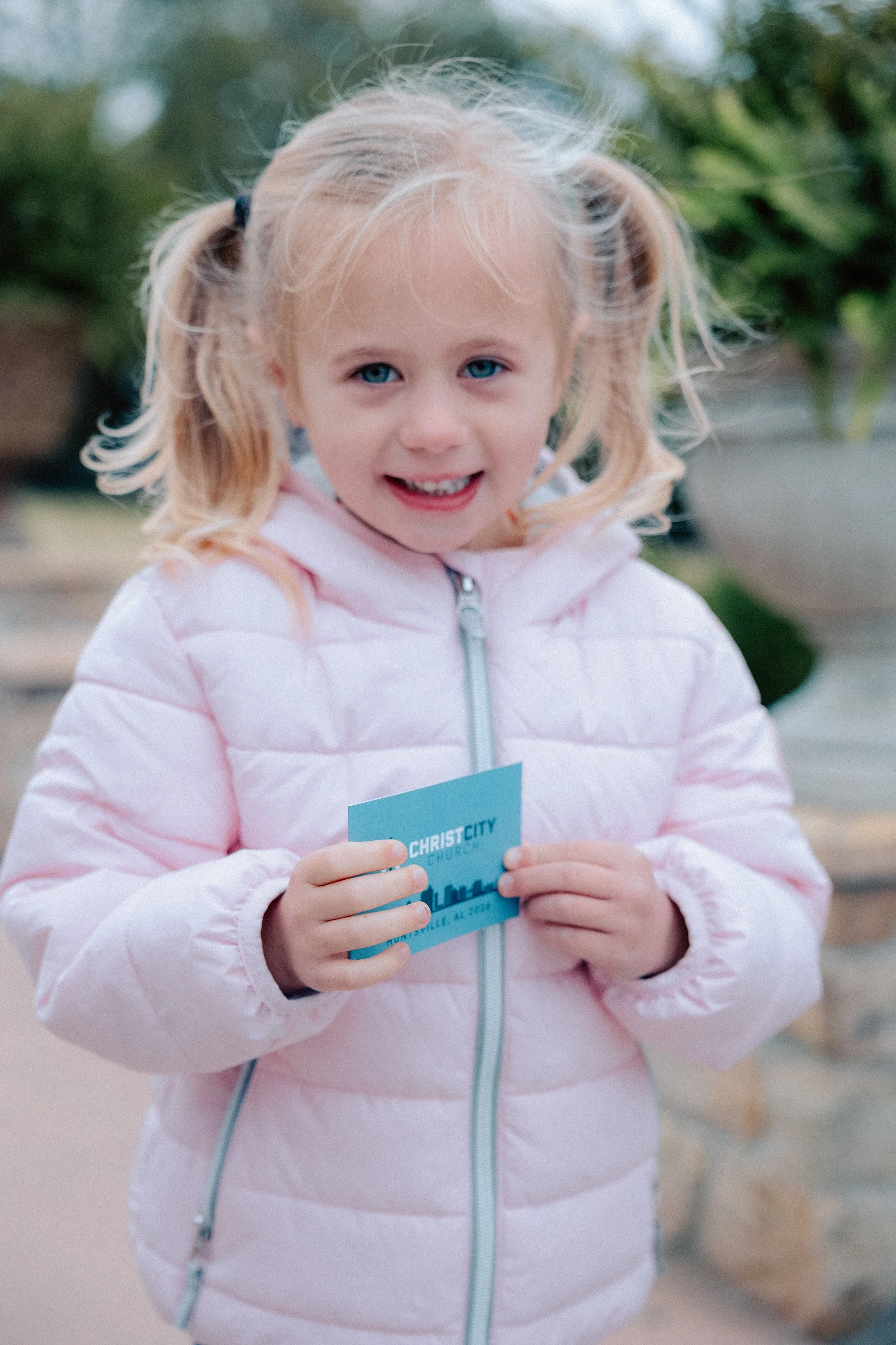 A young girl with blonde hair in pigtails, smiling and holding a blue card from Christ City Church outdoors.