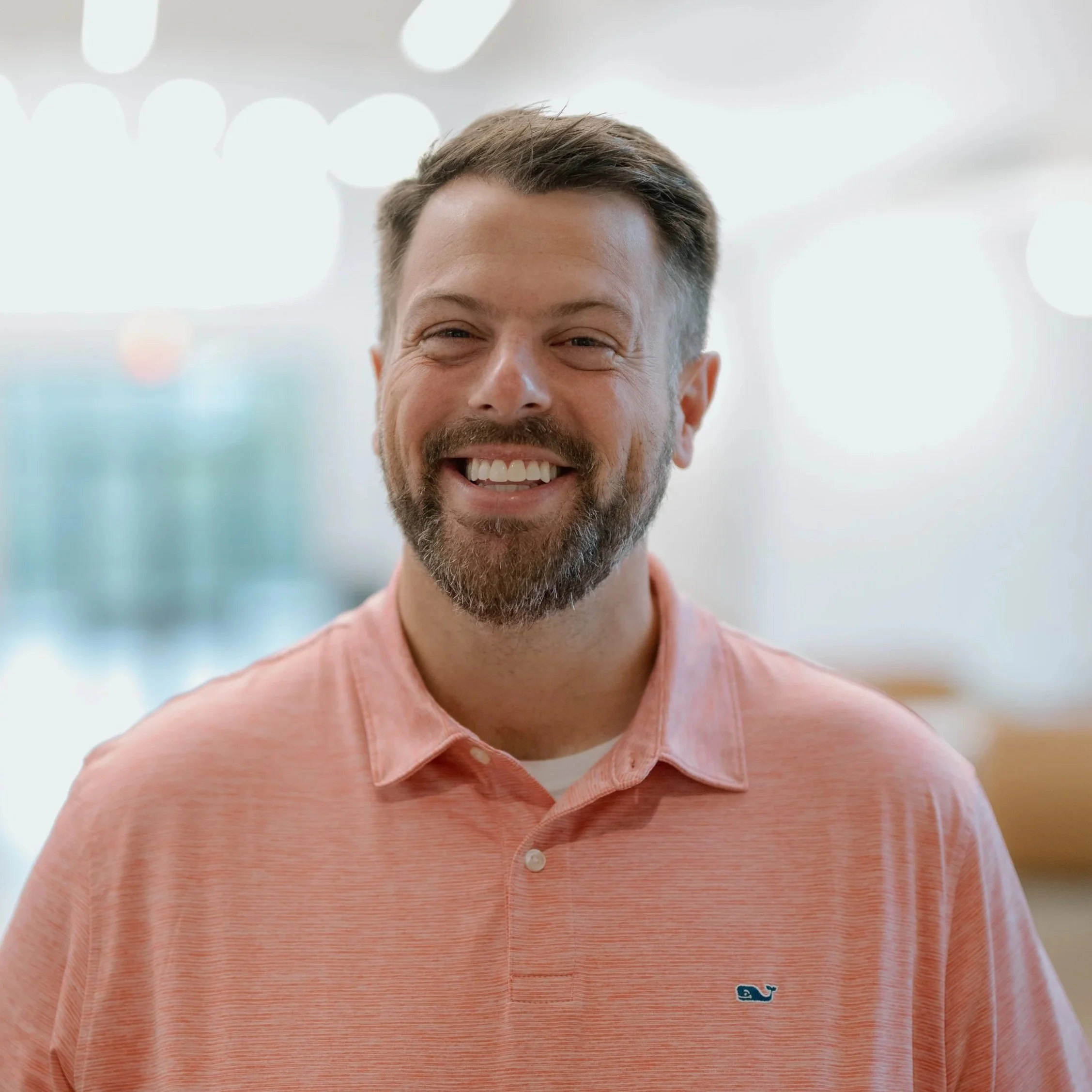 A smiling man standing outdoors in front of trees, wearing a blue button-up shirt.
