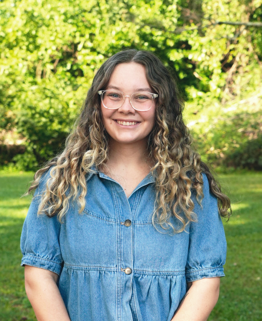 Young woman with curly blond hair smiling outdoors in front of green leafy trees.