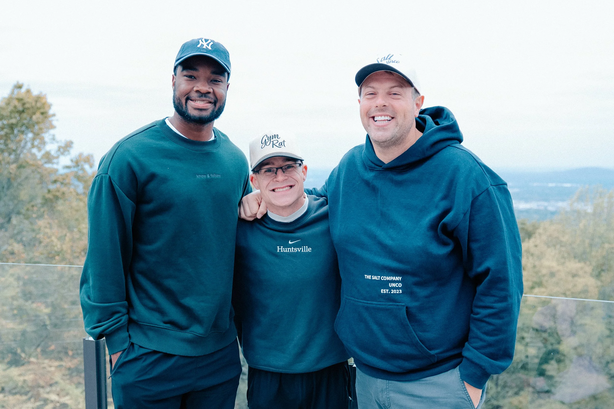 Three men smiling outdoors in casual clothing, standing close together with arms around each other's shoulders, overlooking a landscape with trees and sky.