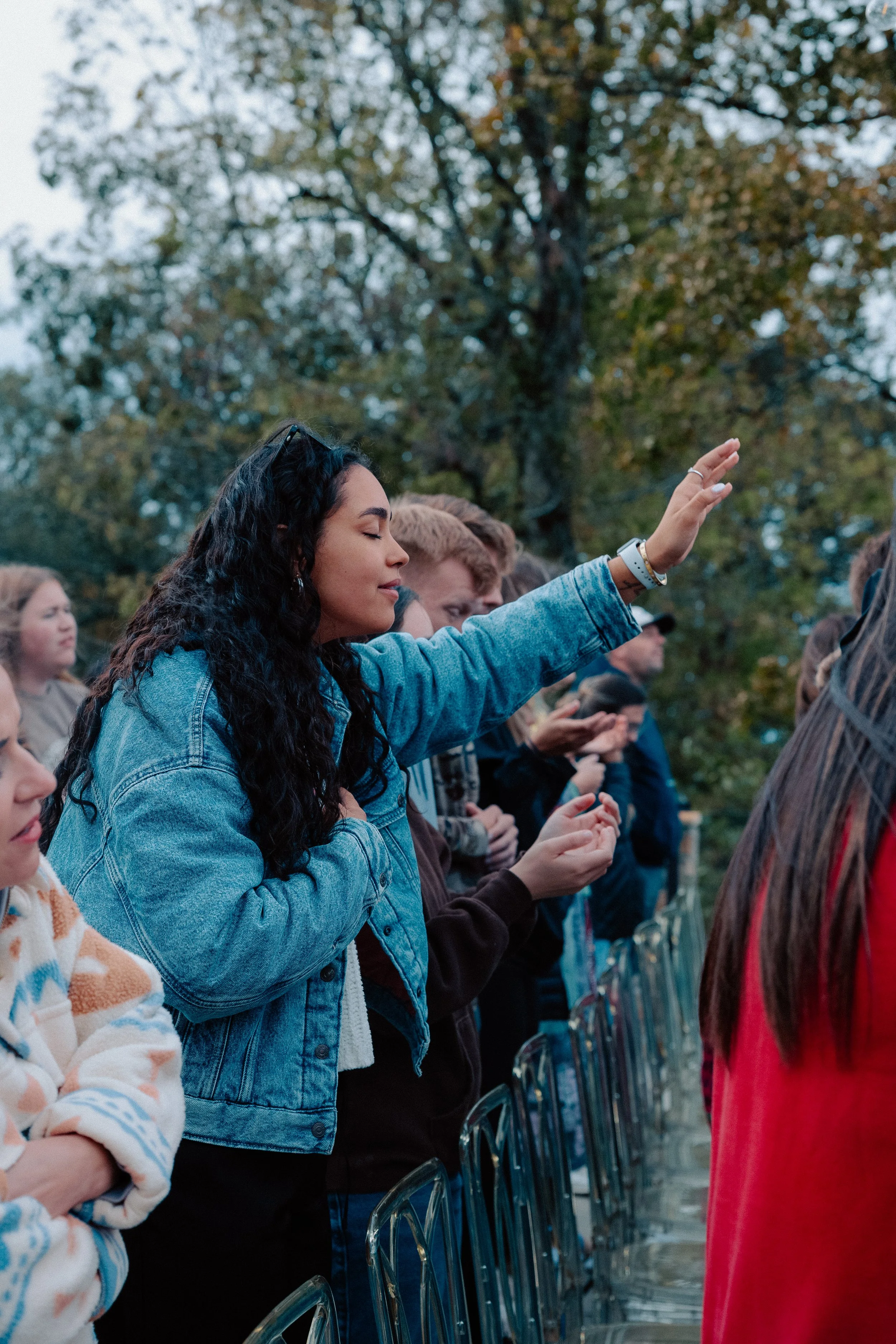 A woman with long, curly black hair wearing a denim jacket stands with her eyes closed and her right hand raised, participating in a prayer or moment of reflection among a group of people outdoors, with trees in the background.