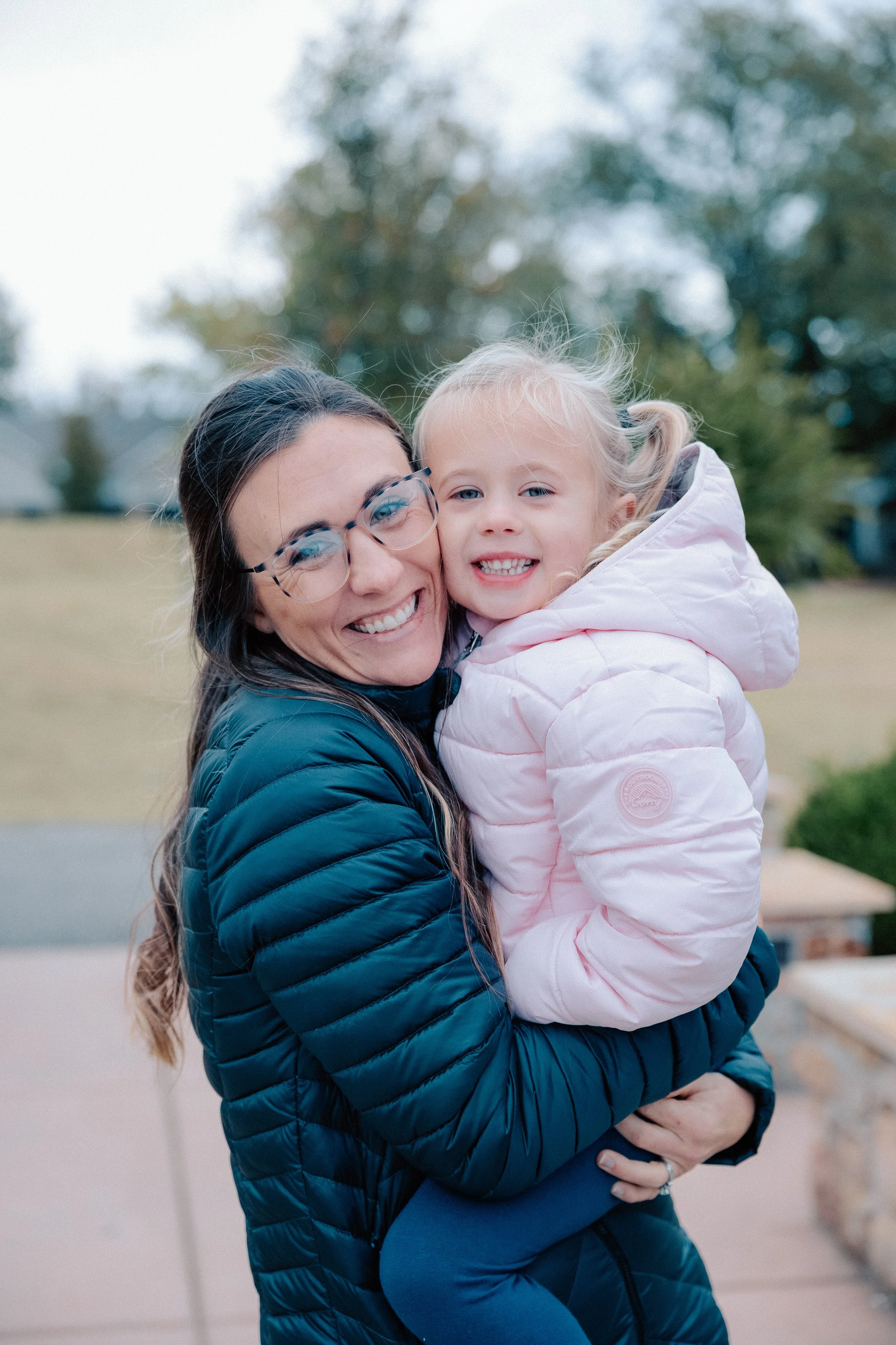 A woman with glasses and a dark blue puffer jacket holding a young girl in a pink puffer jacket outdoors, both smiling.