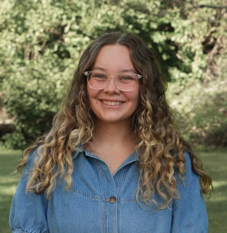 Young woman with curly blond hair smiling outdoors in front of green leafy trees.