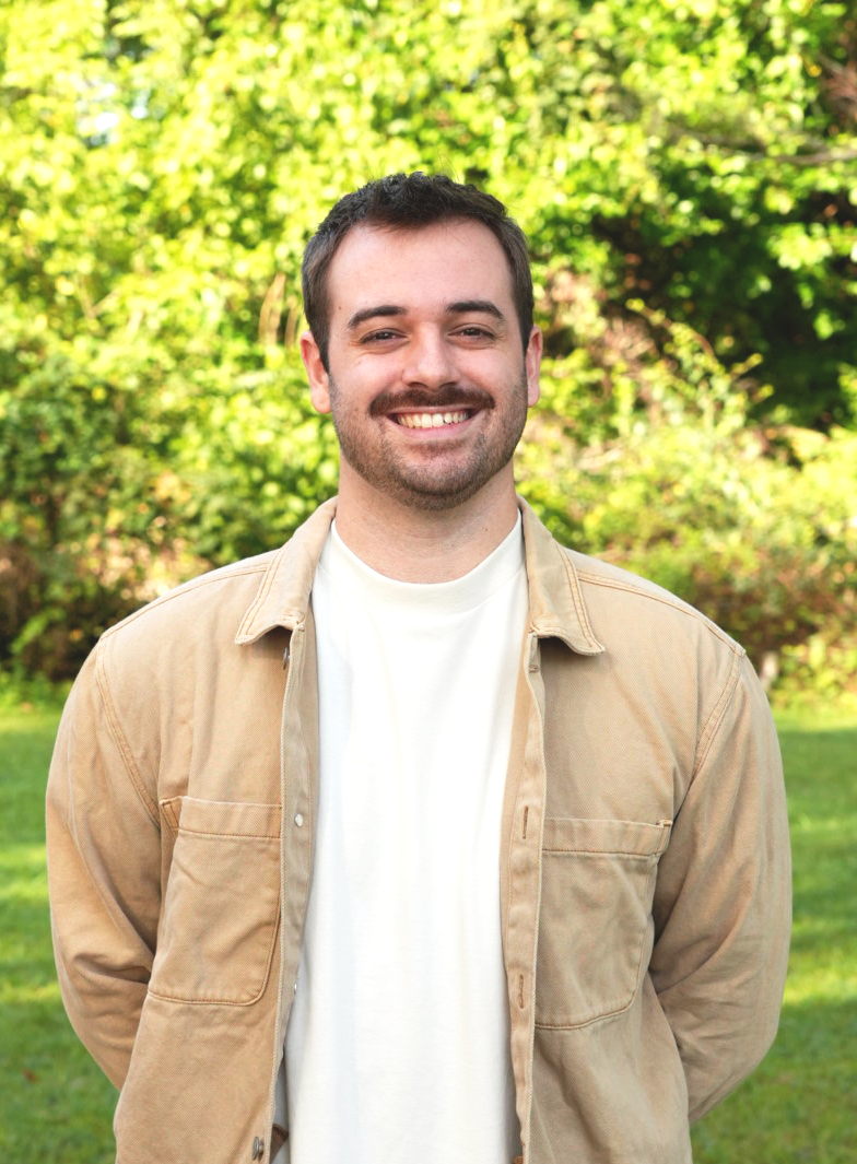 A man with a beard and short hair smiling, standing outdoors in front of green foliage, wearing a light-colored short-sleeve shirt.