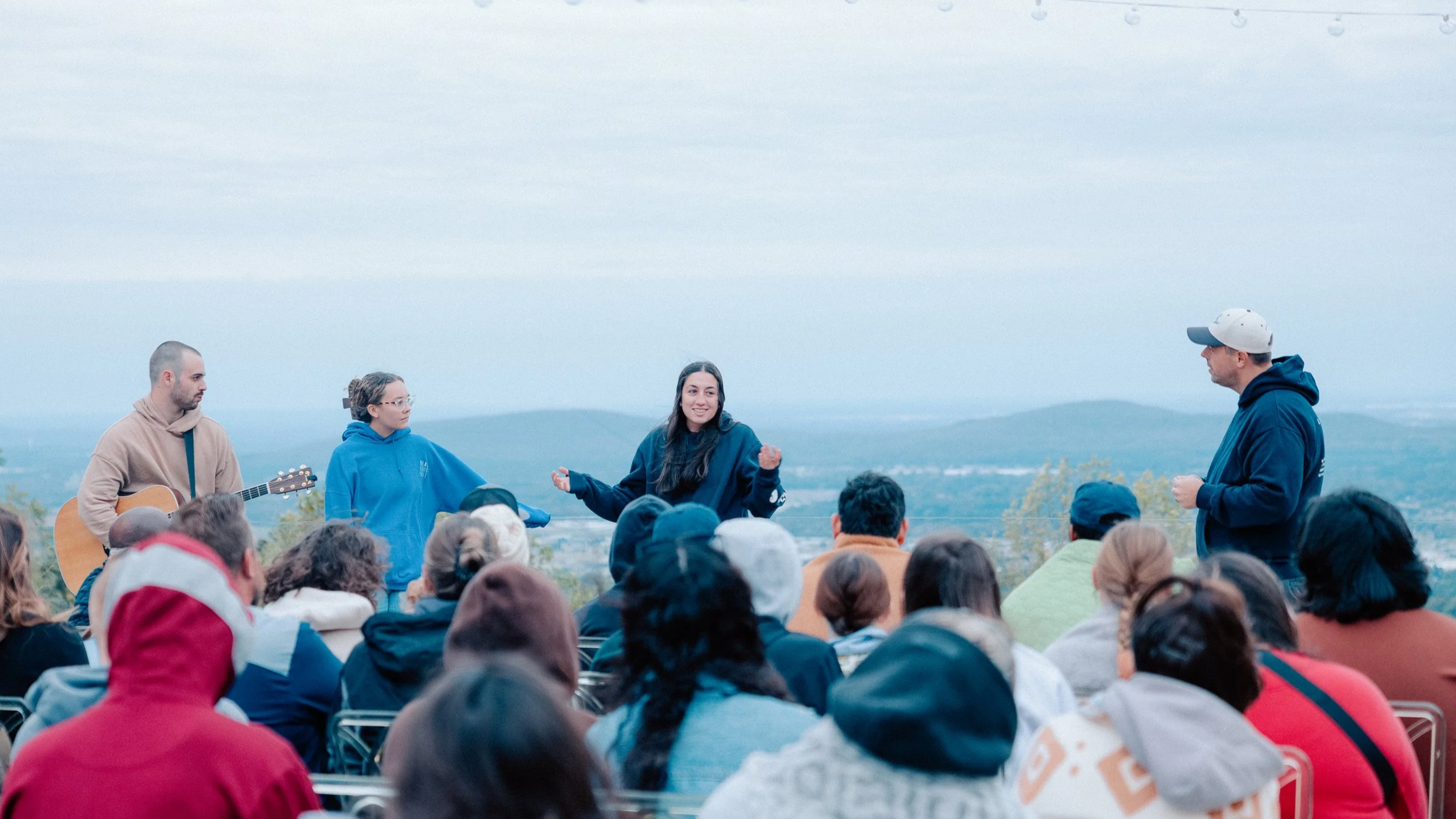 Group of people gathered outdoors with four individuals on stage, one playing a guitar and another speaking, with an audience listening against a backdrop of hills and trees.