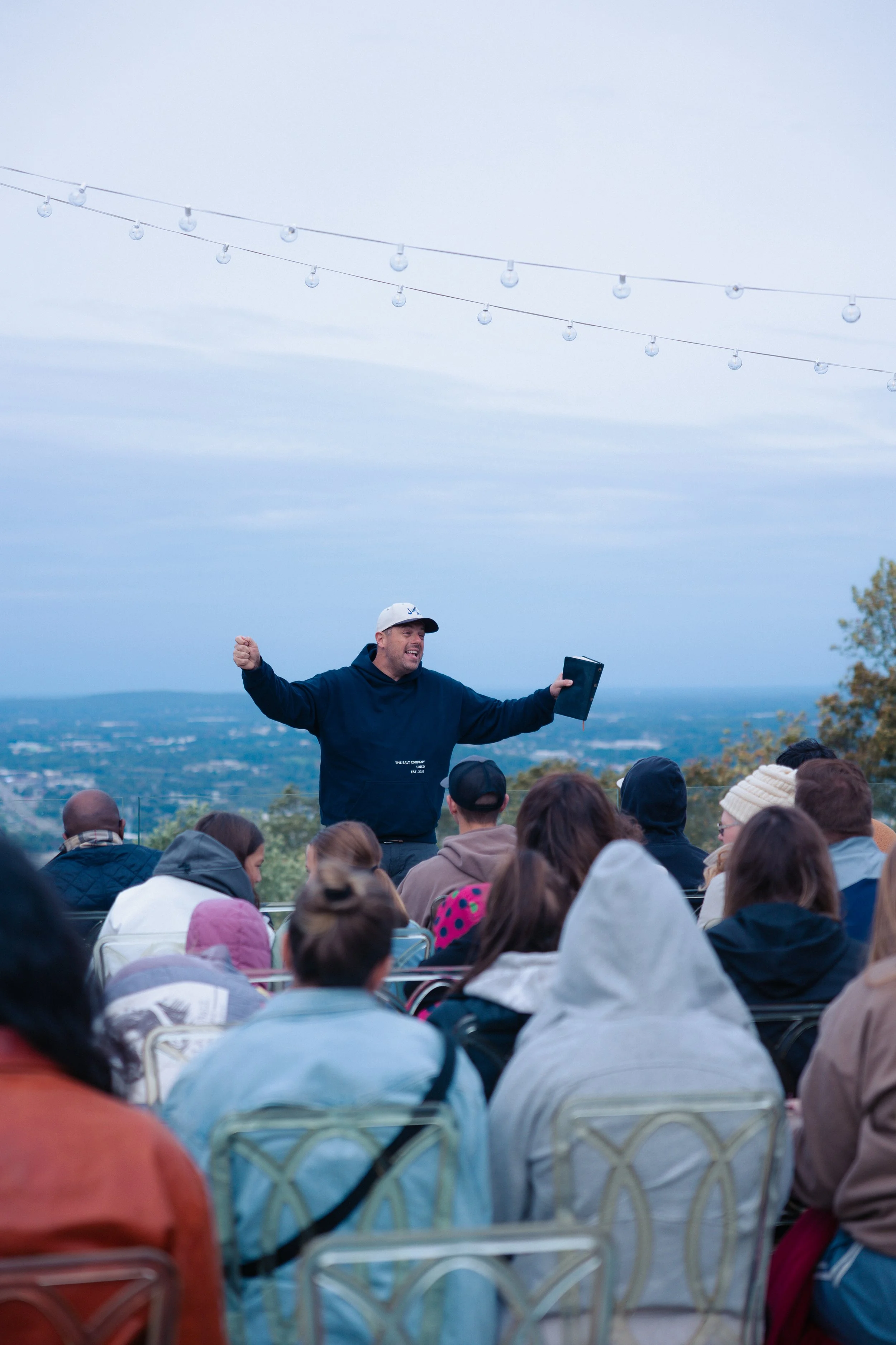 A man in a black hoodie and white cap speaking to an outdoor audience with a scenic view in the background, during an event with string lights overhead.