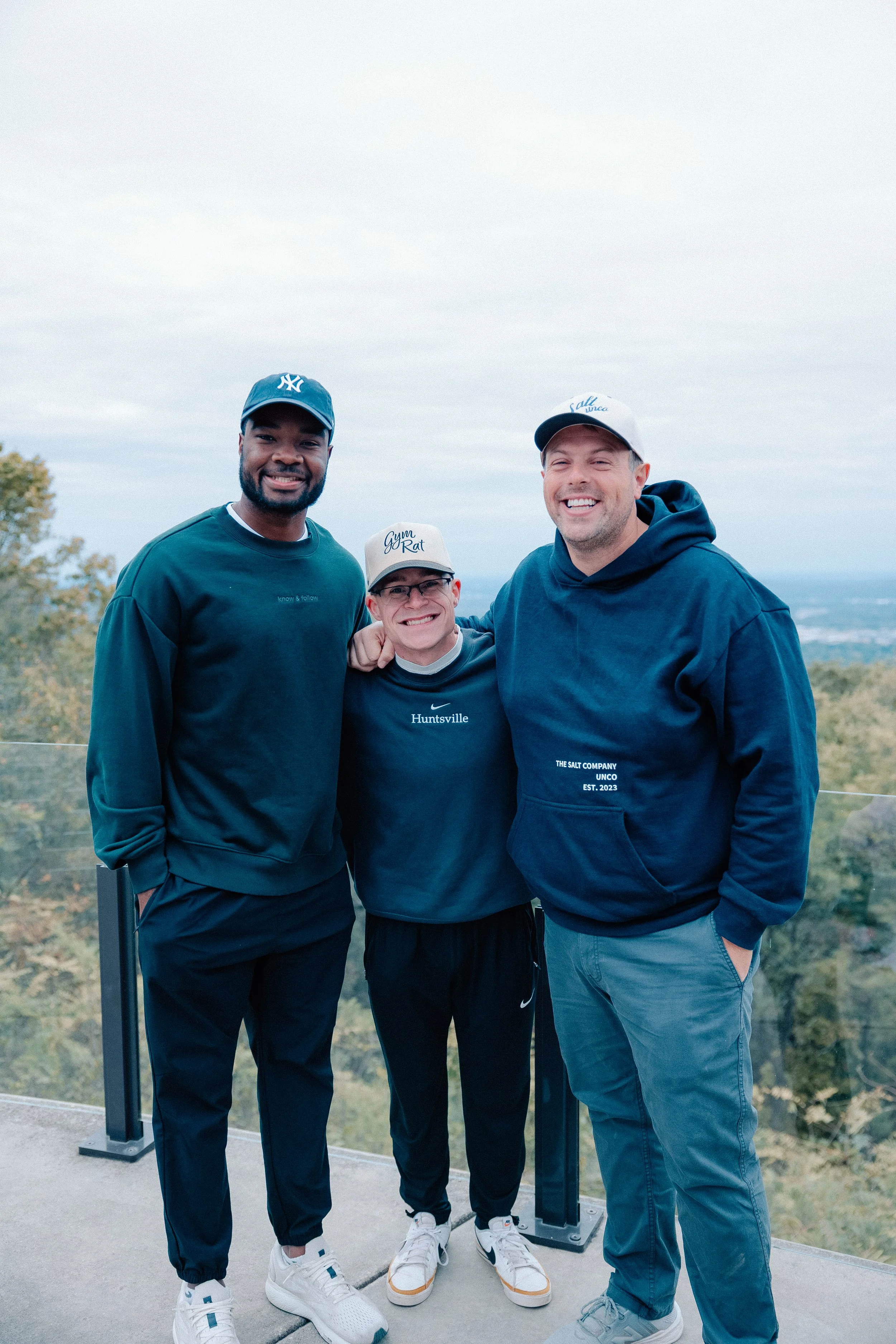 Three men smiling outdoors on a balcony overlooking trees and sky, wearing casual clothing and baseball caps.