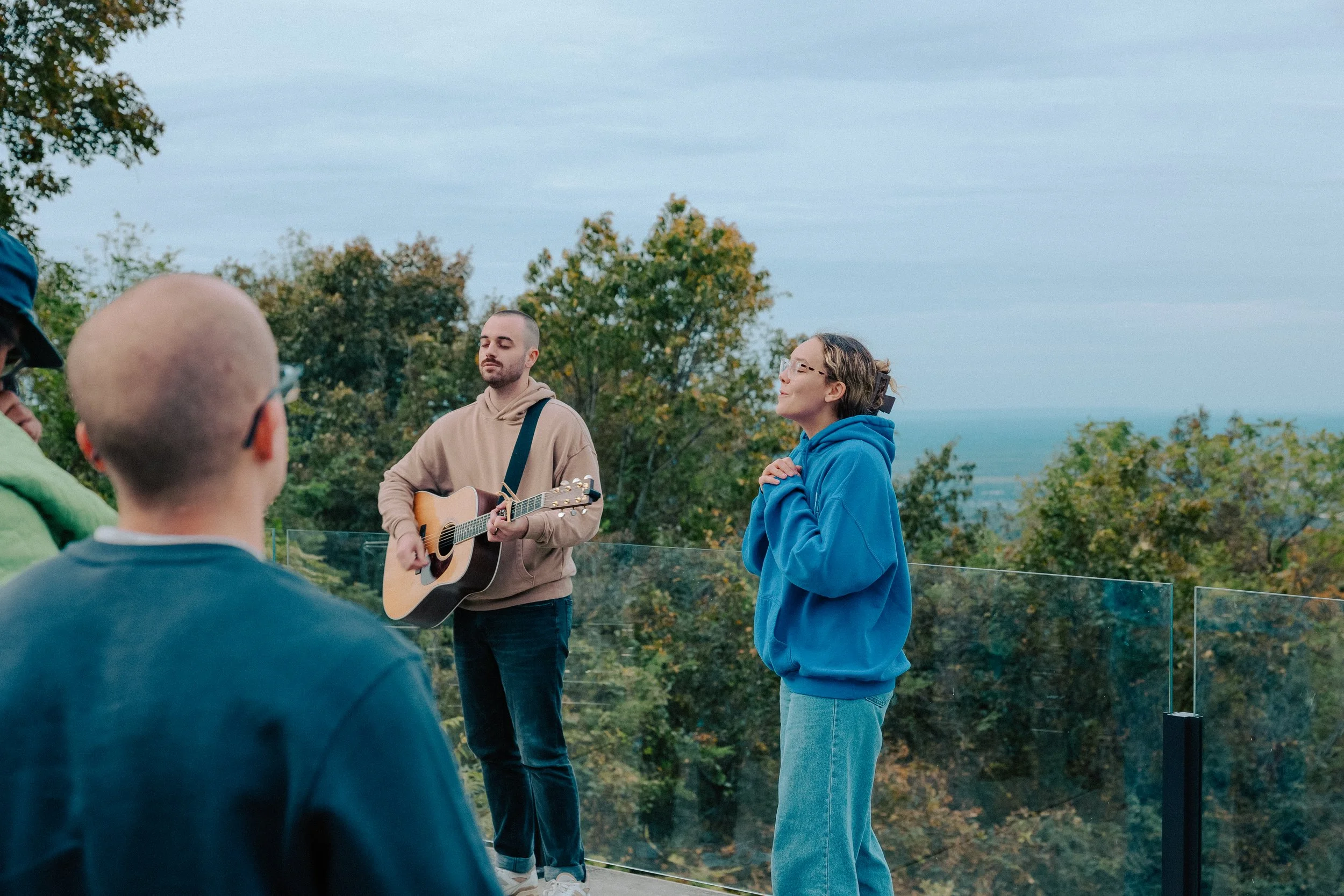 People gathered outdoors on a balcony with a glass railing, enjoying a live acoustic music performance by a man playing an acoustic guitar, while a woman stands next to him with her hands clasped near her chest, all surrounded by trees with a view of
