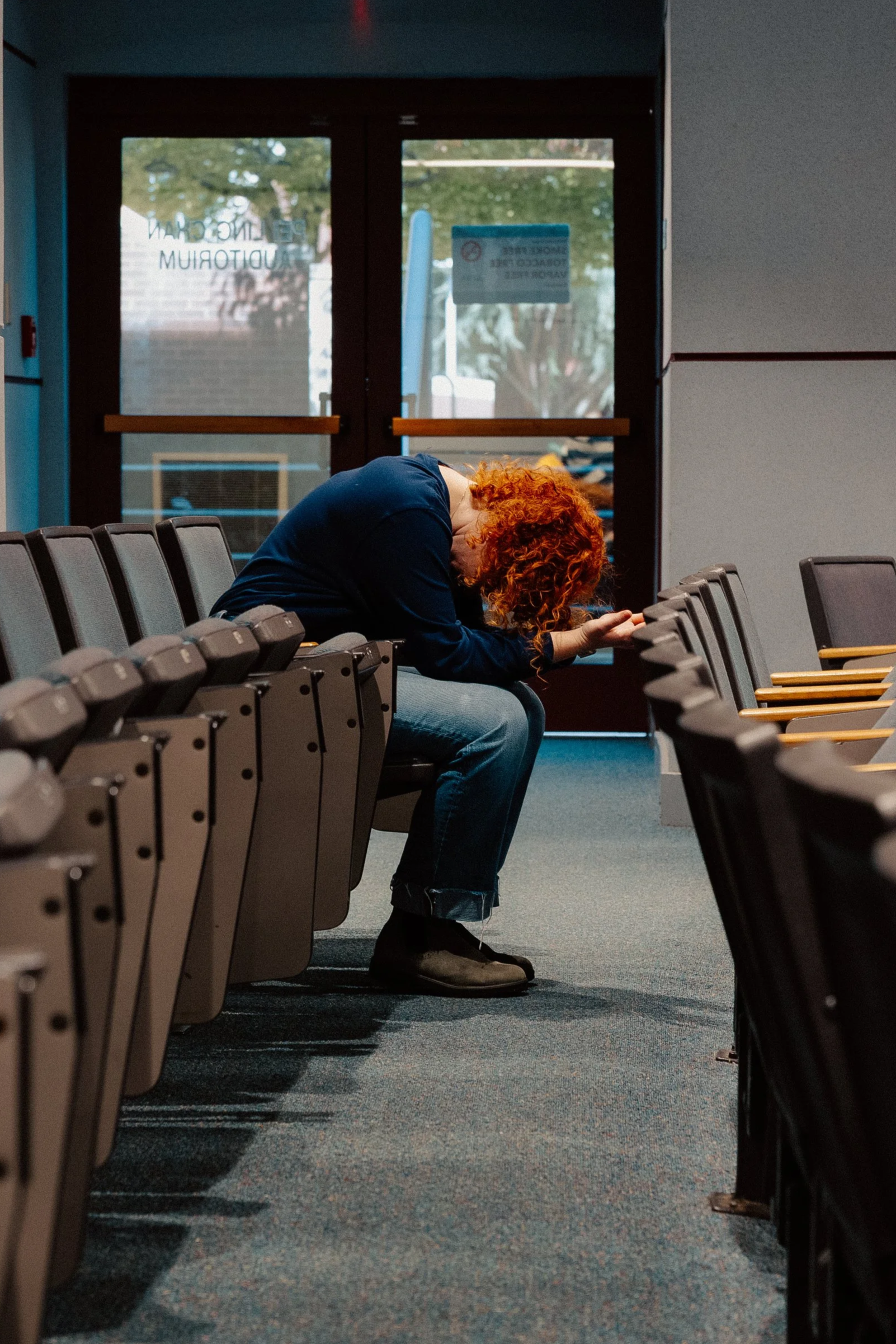 A woman with curly red hair sitting alone in an empty waiting room, head bowed and hands clasped.