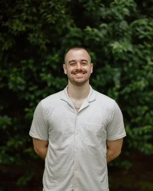 A man with a beard and short hair smiling, standing outdoors in front of green foliage, wearing a light-colored short-sleeve shirt.