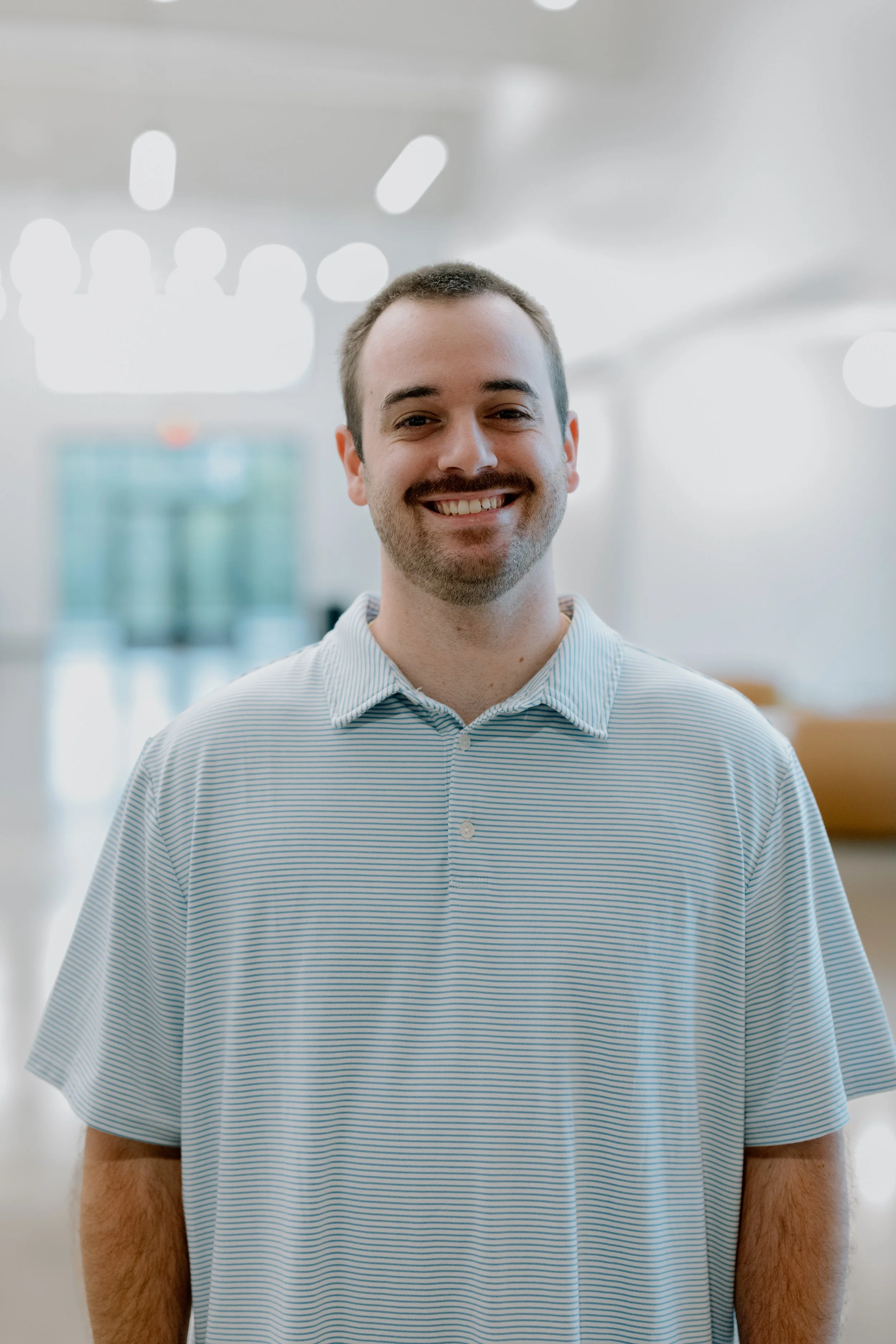 A man with a beard and short hair smiling, standing outdoors in front of green foliage, wearing a light-colored short-sleeve shirt.