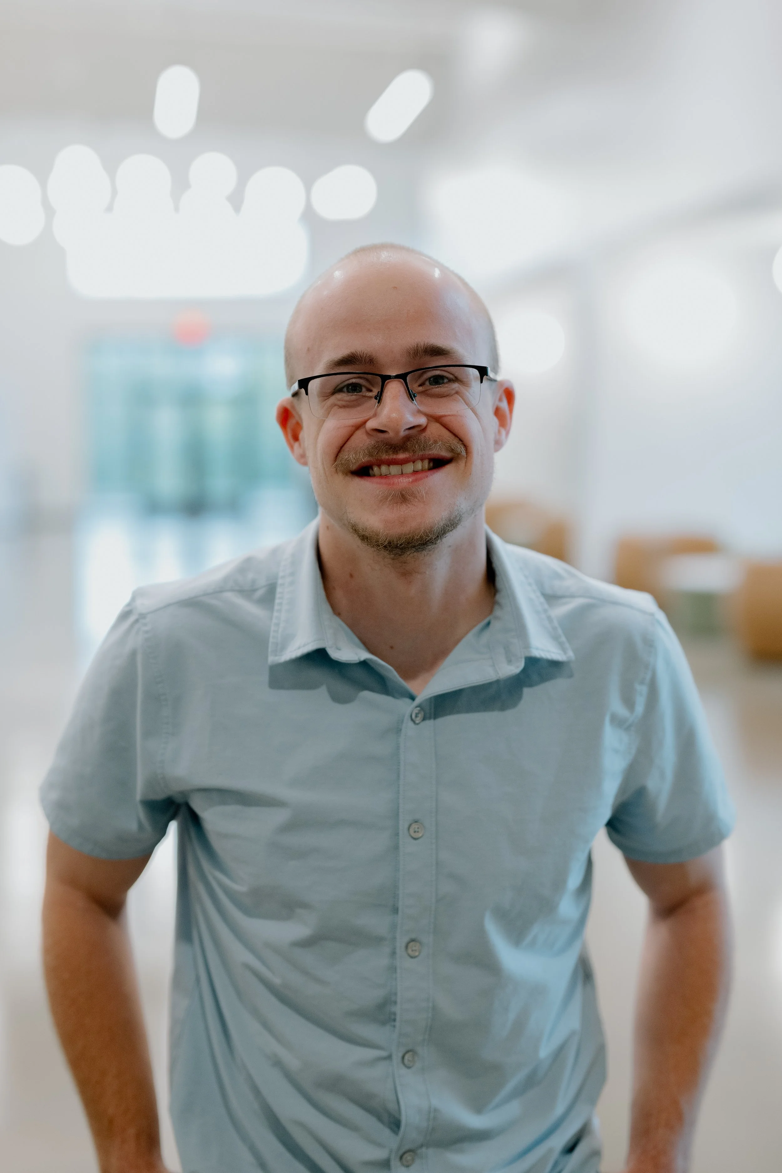 A smiling man wearing glasses and a light blue polo shirt standing outdoors in front of green foliage.