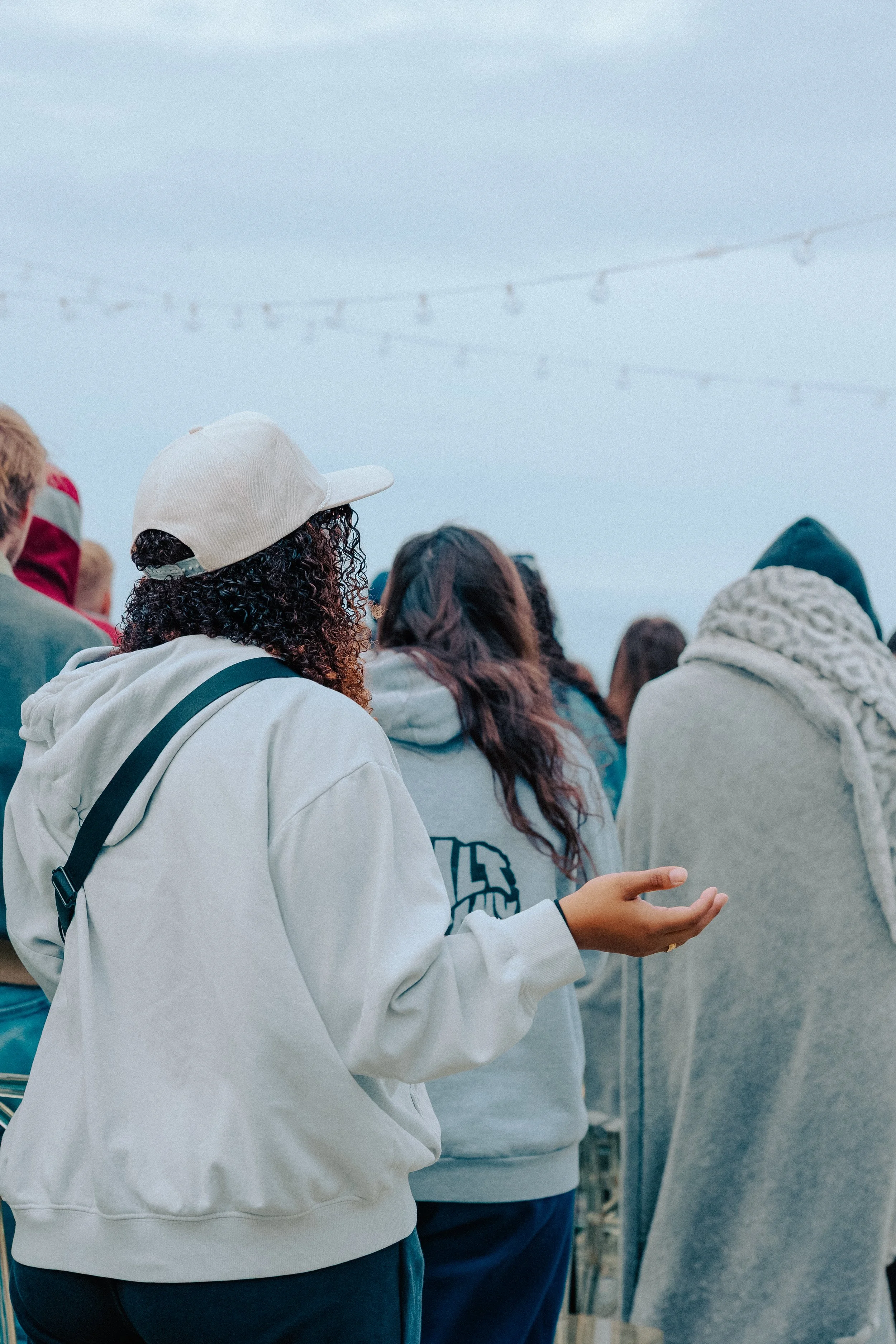 A group of people gathered outdoors during daytime, facing away from the camera, with some wearing hoodies and jackets, under a cloudy sky with string lights overhead.