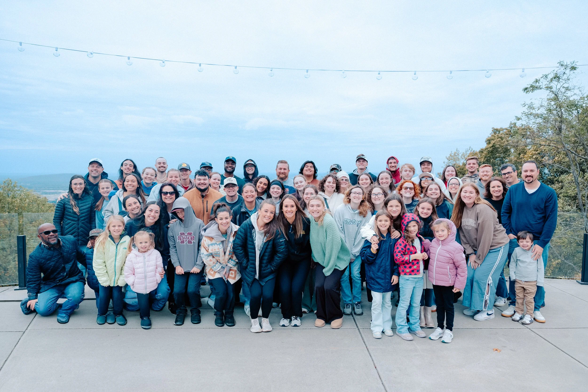 Large group of people gathered outdoors on a cloudy day, posing for a photo on a concrete surface, with trees and a faint landscape in the background.