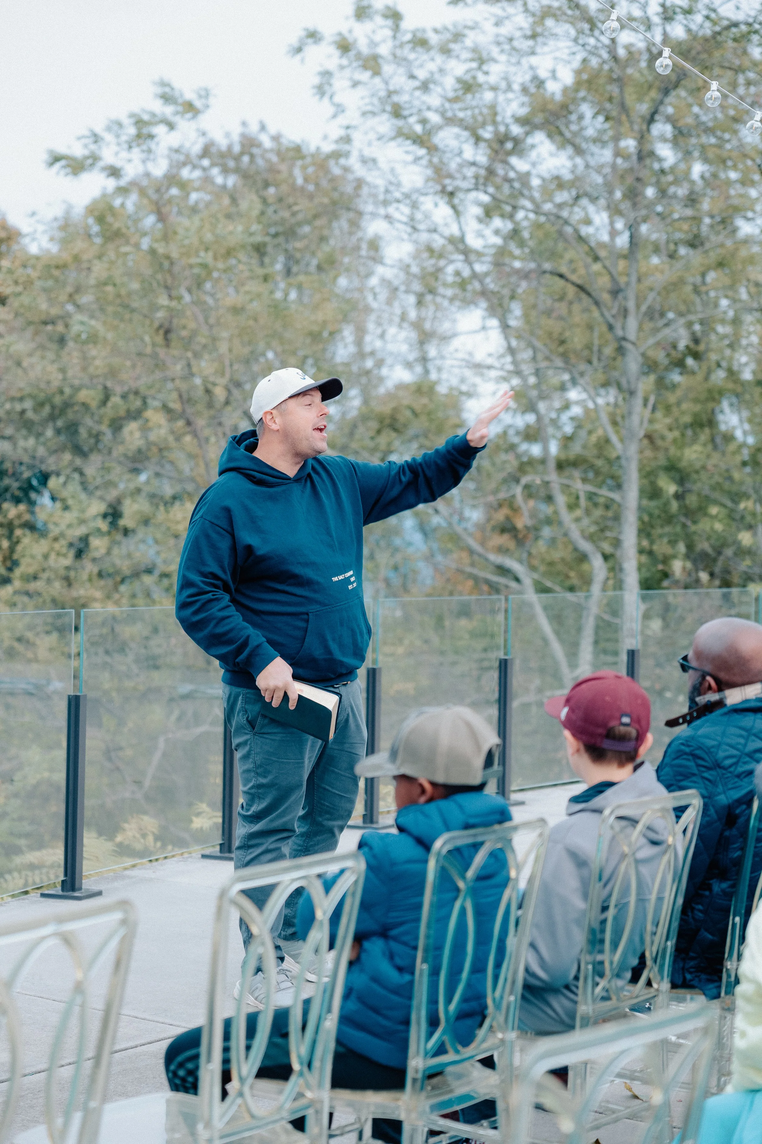A man in a blue hoodie and baseball cap is speaking to a group of children seated outdoors, with trees and string lights in the background.