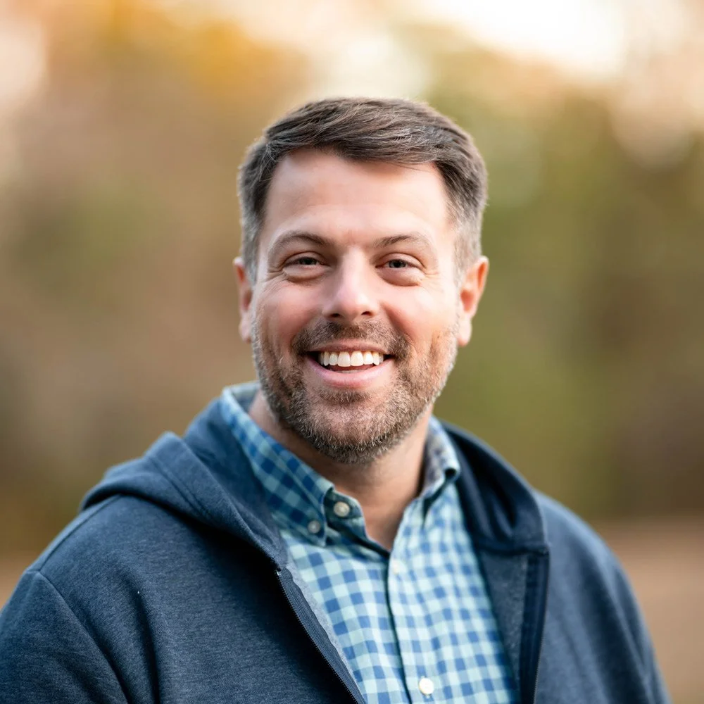 A smiling man standing outdoors in front of trees, wearing a blue button-up shirt.