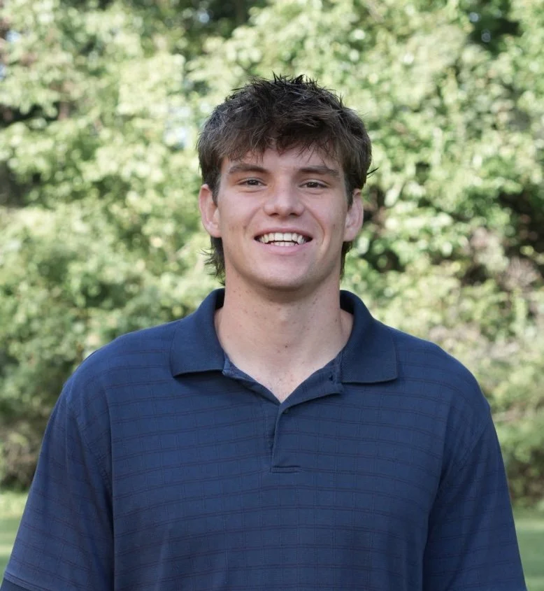 Young man smiling outdoors with green trees in the background.