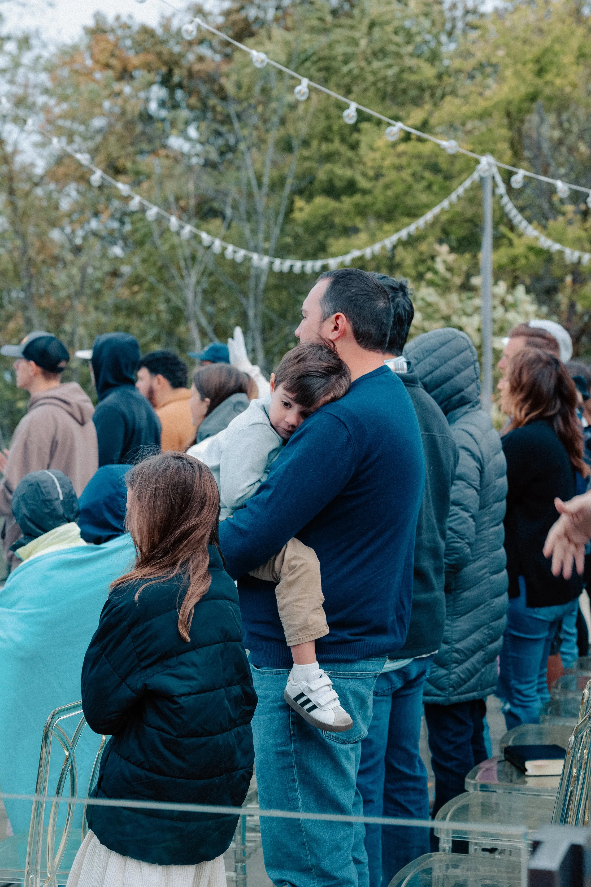 A man holding a young boy in a crowd during an outdoor event with trees and string lights in the background.
