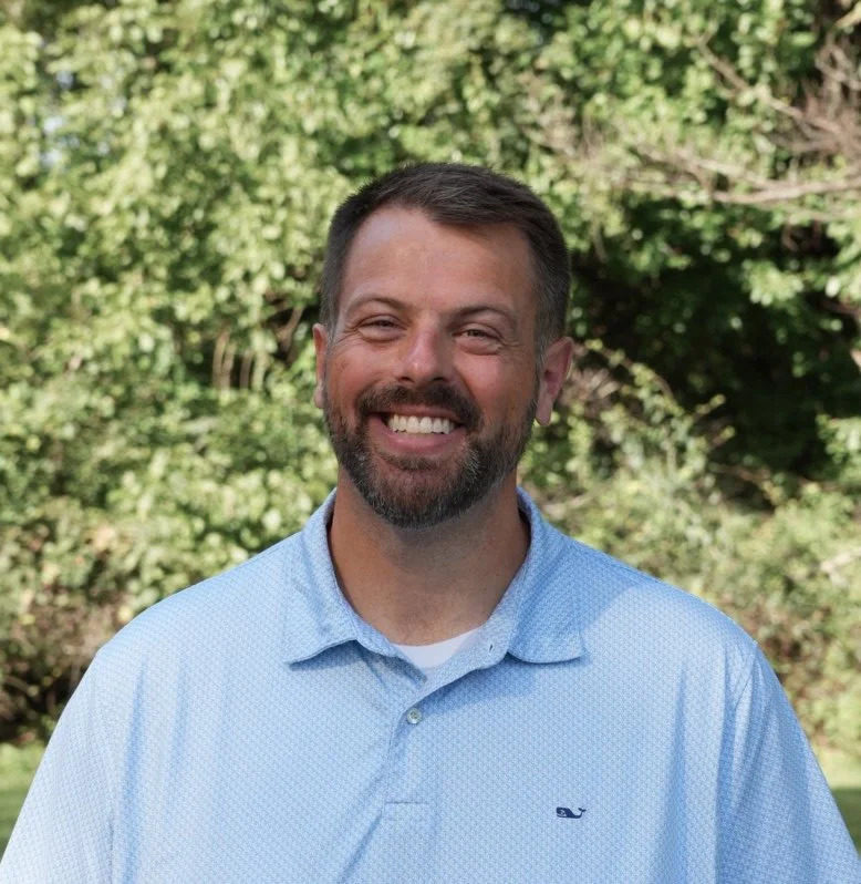 A smiling man standing outdoors in front of trees, wearing a blue button-up shirt.